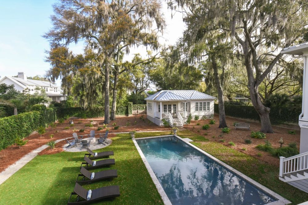 Elevated view of a landscaped backyard featuring a rectangular swimming pool, lounge area, fire pit, and white guest house under oak trees with Spanish moss