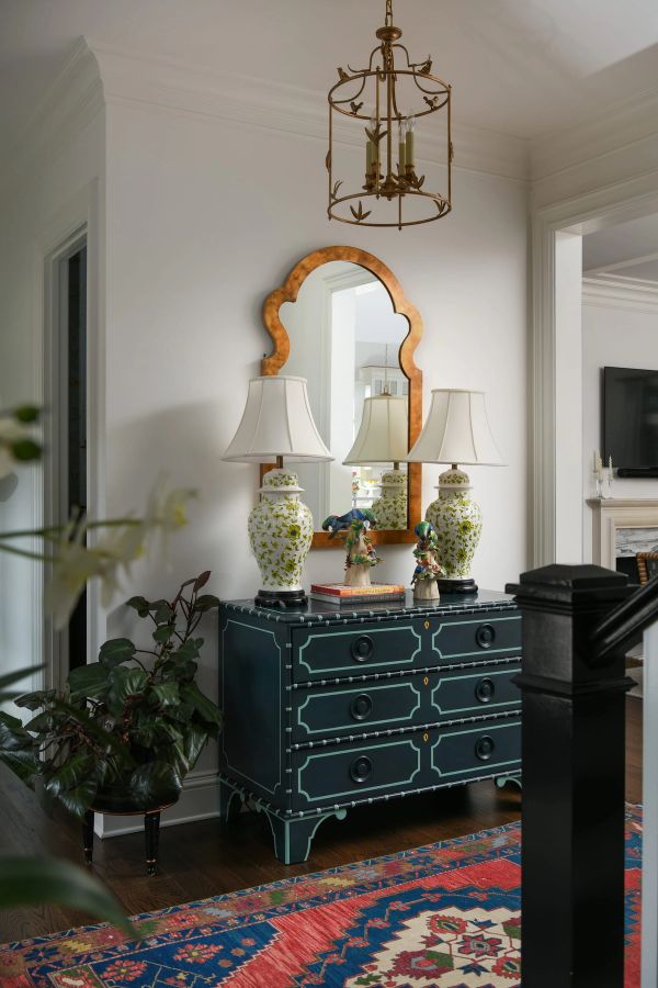 Elegant hallway entry featuring a dark teal chest of drawers, scalloped gold mirror, gold birdcage chandelier, and vibrant patterned rug