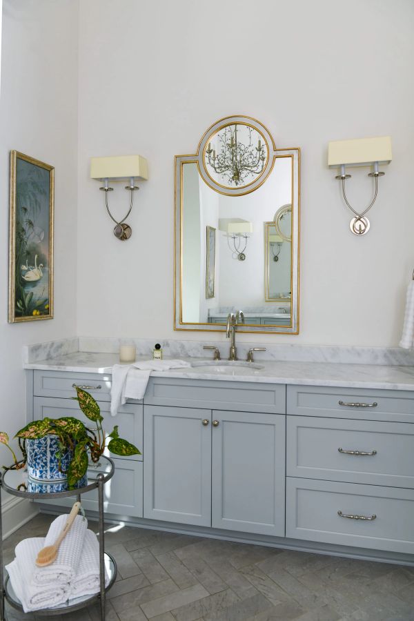Bathroom vanity detail with pale blue-gray cabinetry, marble countertop, gold-framed mirror, wall sconces, and a rolling cart with towels and greenery