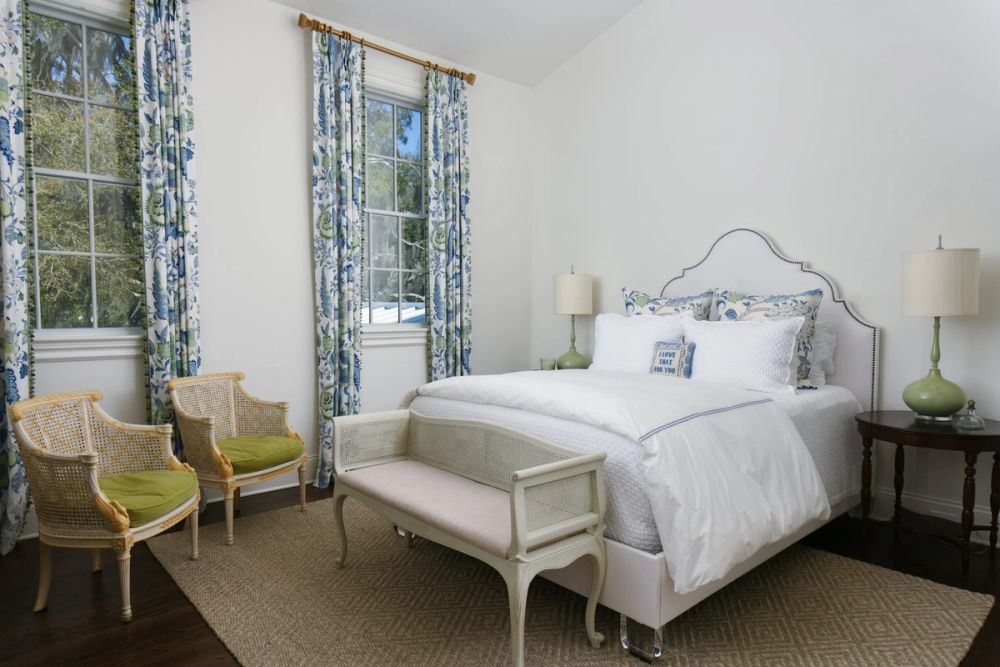 Bright guest bedroom with a white upholstered bed, blue and green floral curtains, green cane-back armchairs, and cane bench at the foot of the bed