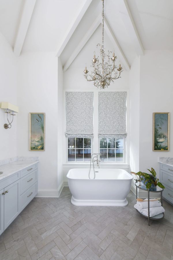 Primary bathroom featuring a freestanding tub beneath a vaulted ceiling, crystal chandelier, swan artwork, and light grey vanities with marble countertops