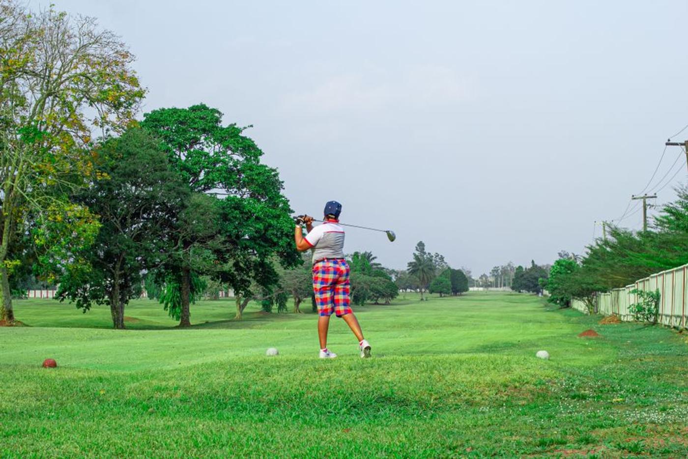 A golfer takes a full swing with a driver on a tree-lined fairway, golf balls resting nearby