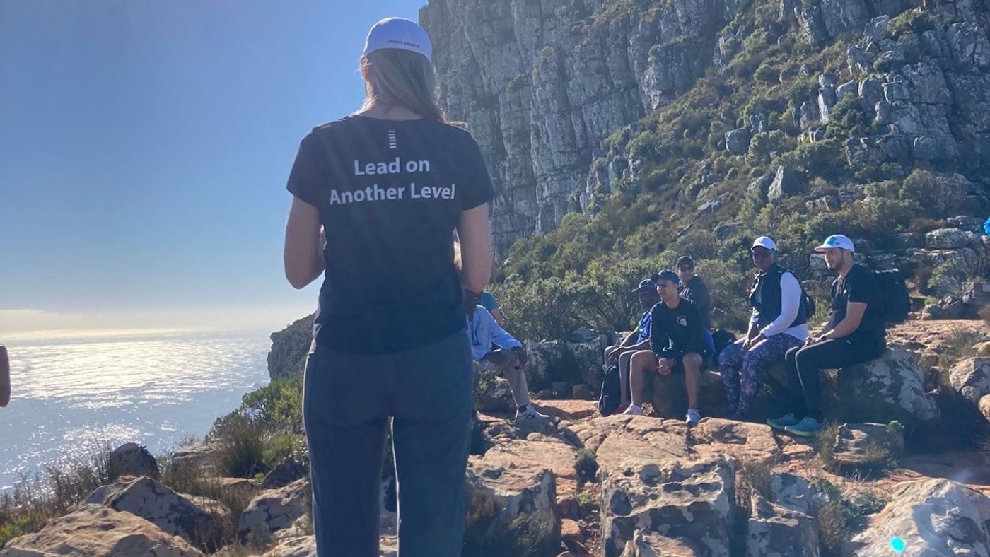 A TLH participant in a “Lead on Another Level” shirt looking out over the sea from a mountainside, with the group resting behind
