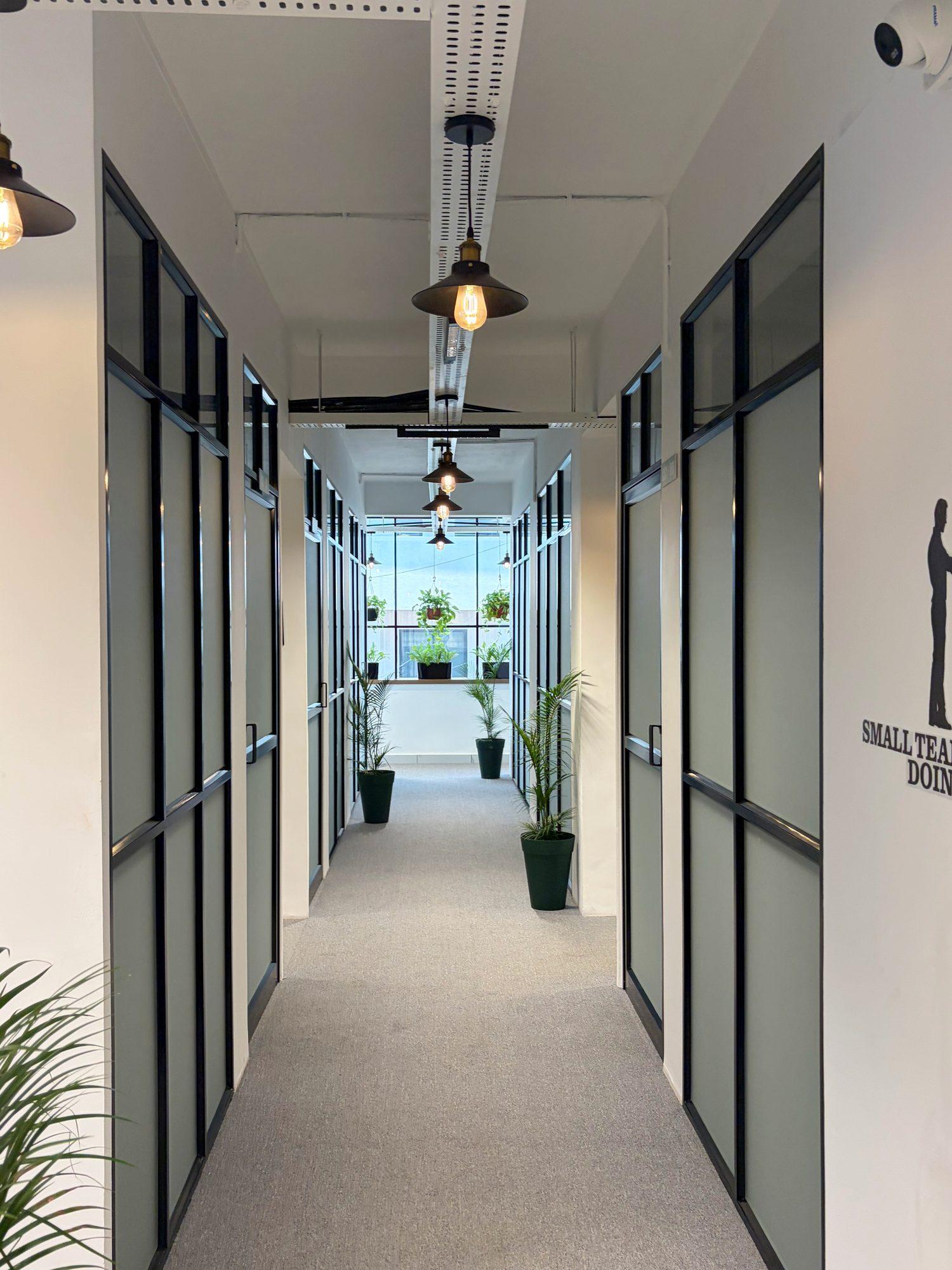 Corridor of glass-panelled cabins with pendant lights and palms leading to a window of hanging plants