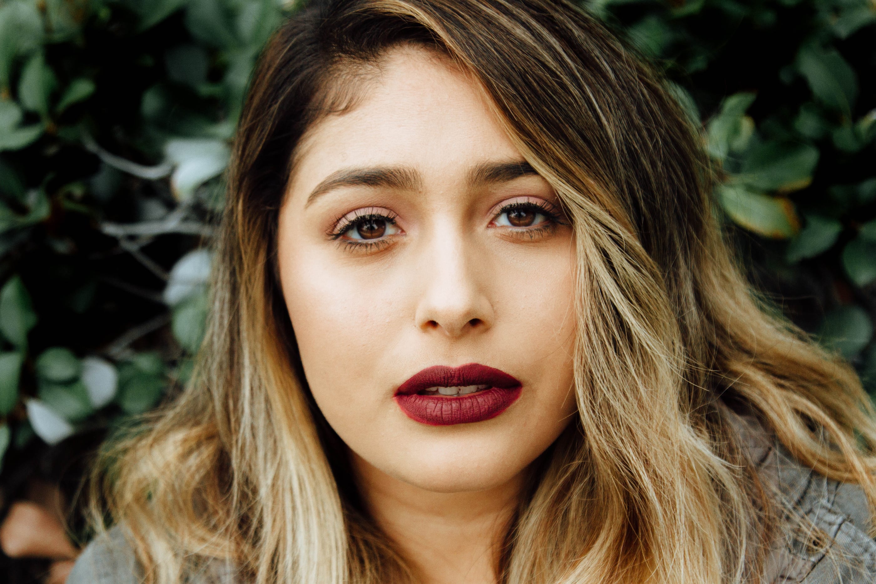 Portrait of a woman with dark red lipstick and long highlighted hair, against green leaves