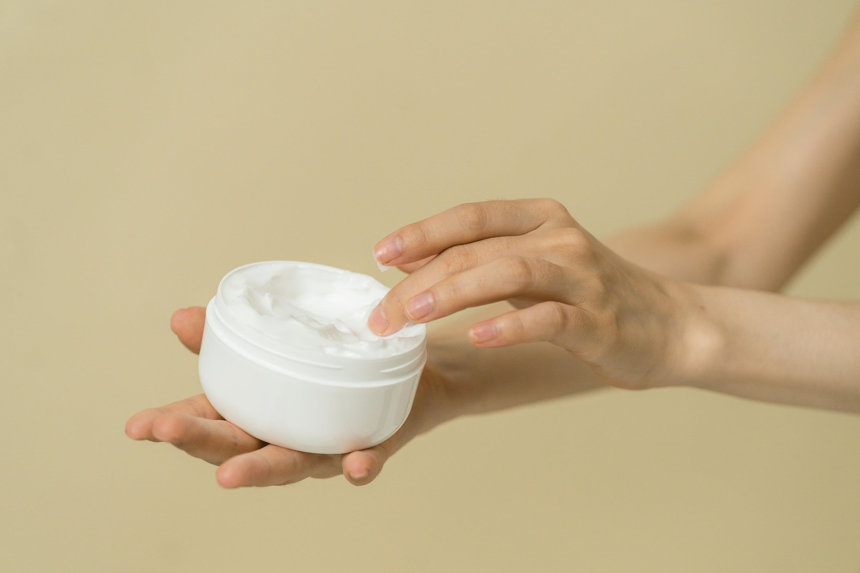 A hand holding an open jar of face cream against a warm beige background