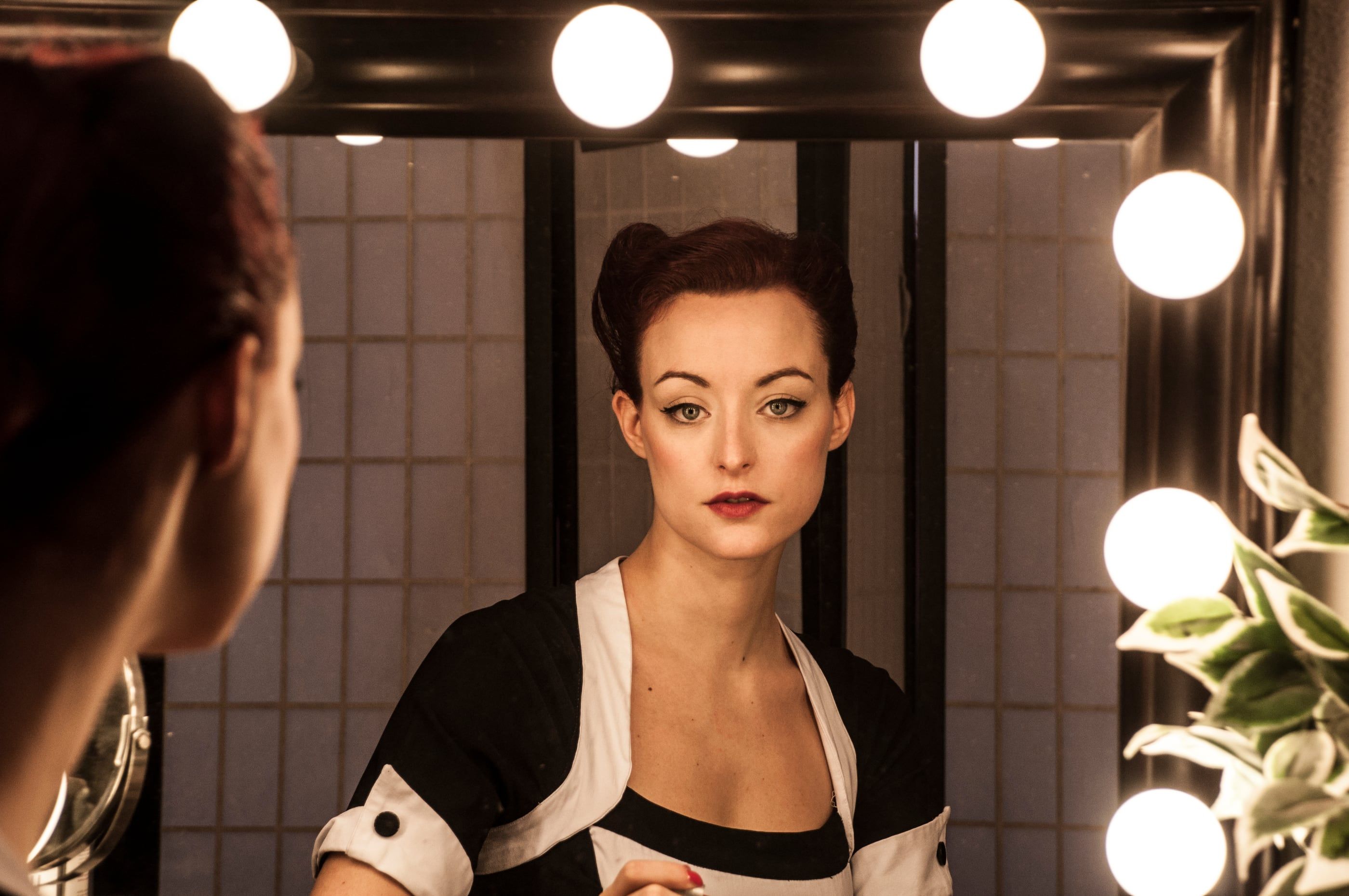 A woman with vintage-styled hair and red lipstick, reflected in a bulb-lit dressing-room mirror