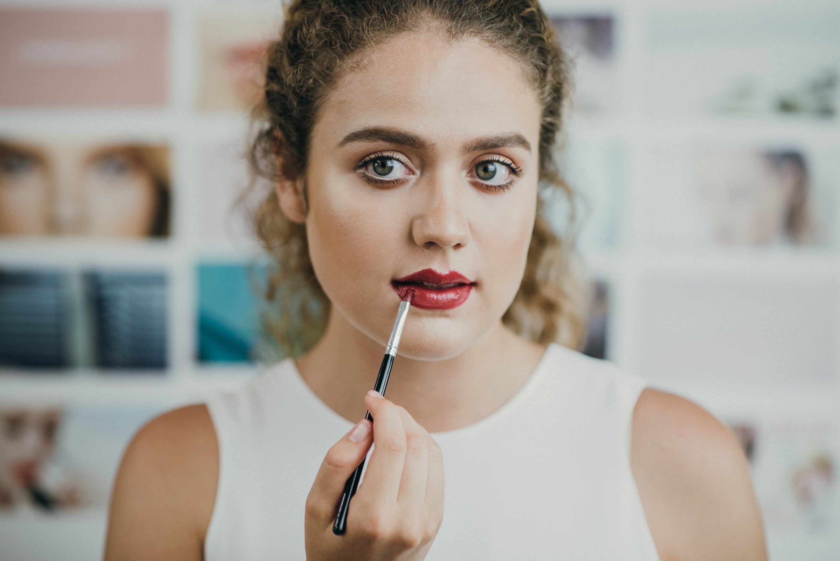 A woman applying red lipstick with a lip brush, in front of a wall of beauty photographs