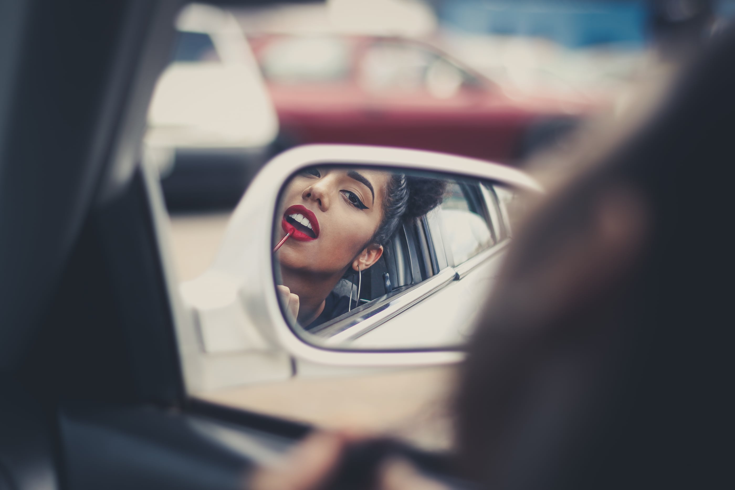 A woman applying red lipstick, reflected in a car's wing mirror