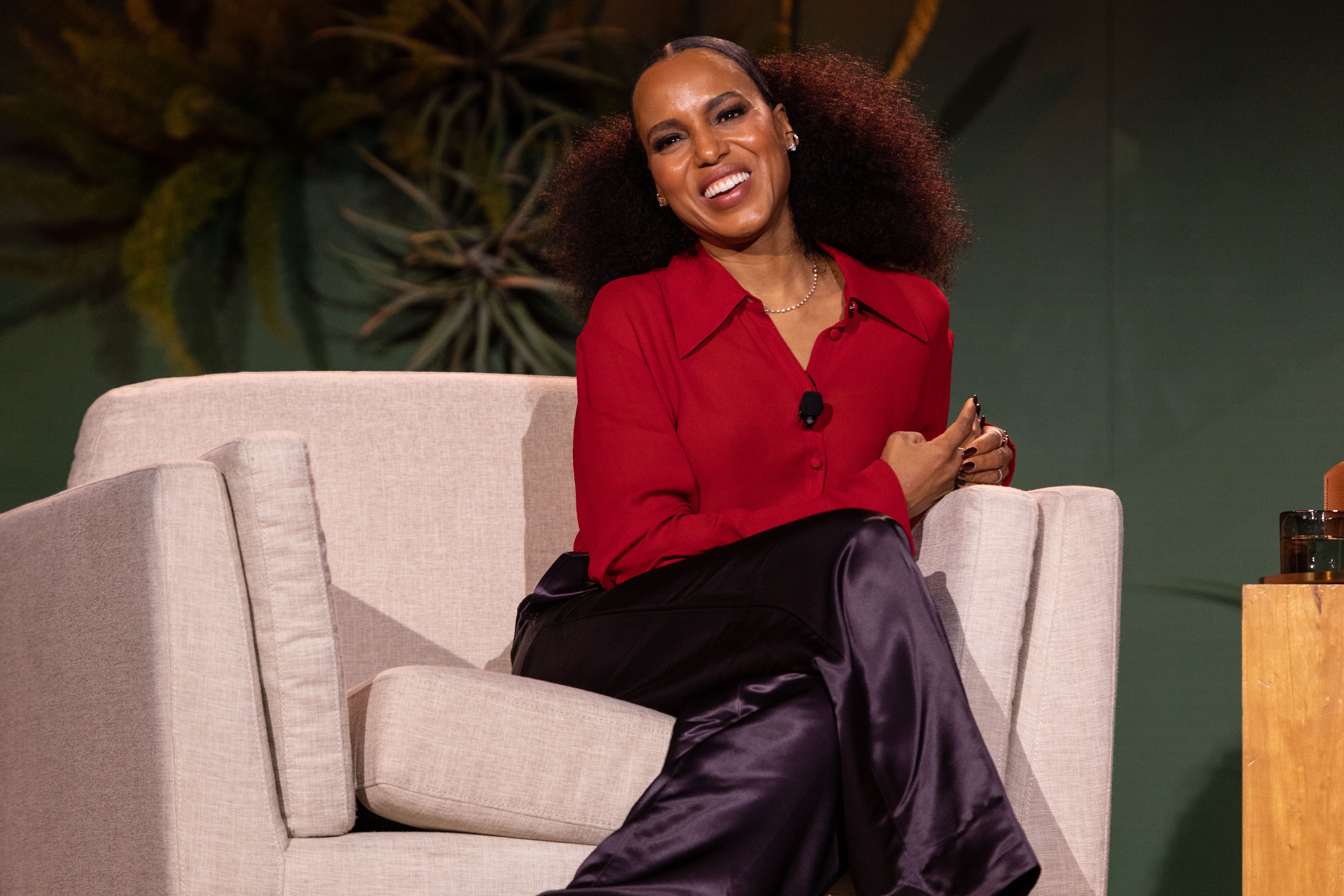 Kerry Washington sitting in a chair smiling with a red shirt and black pants. 