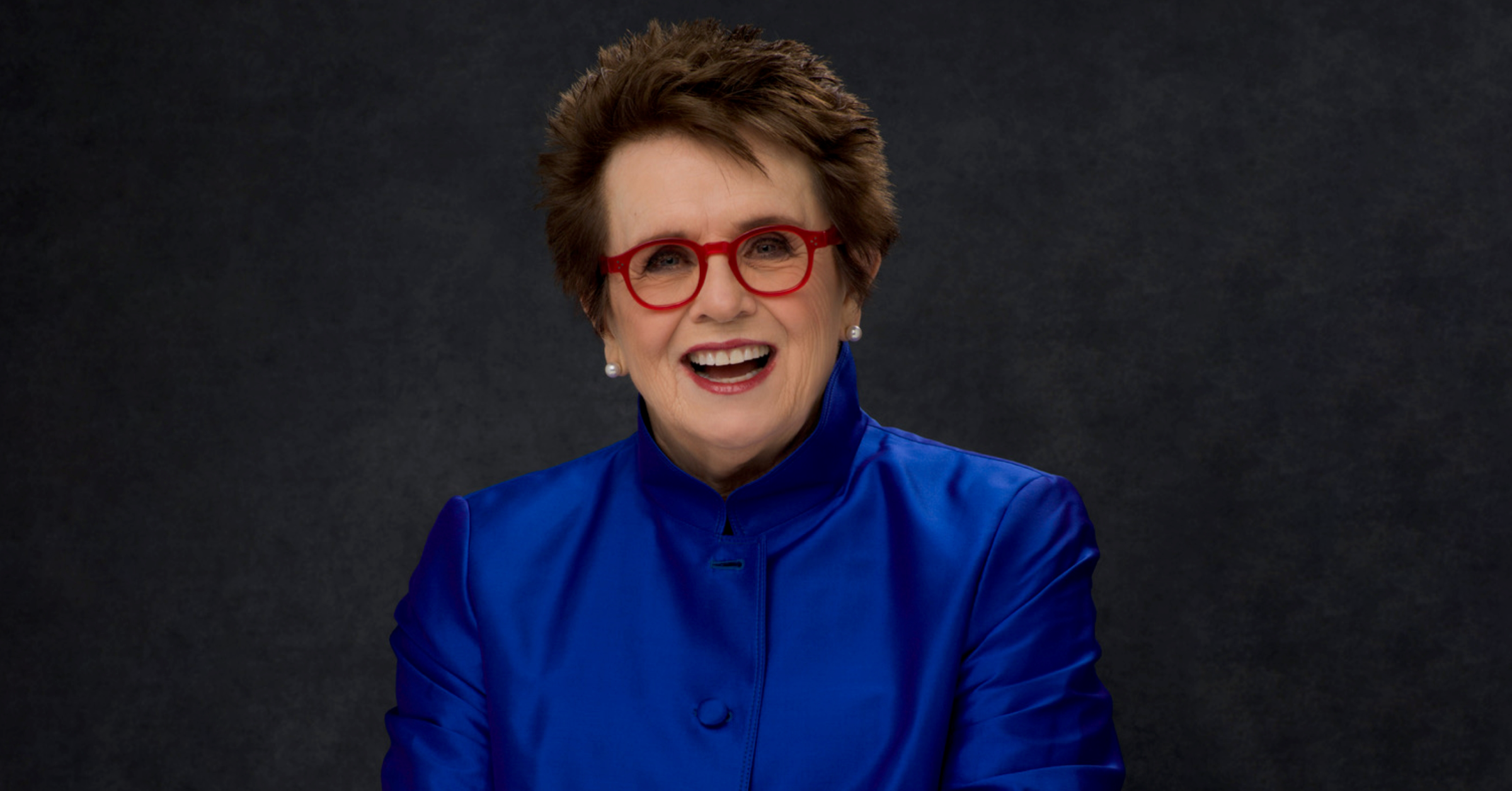 Billie Jean King smiling in a blue blazer and wearing red glasses.