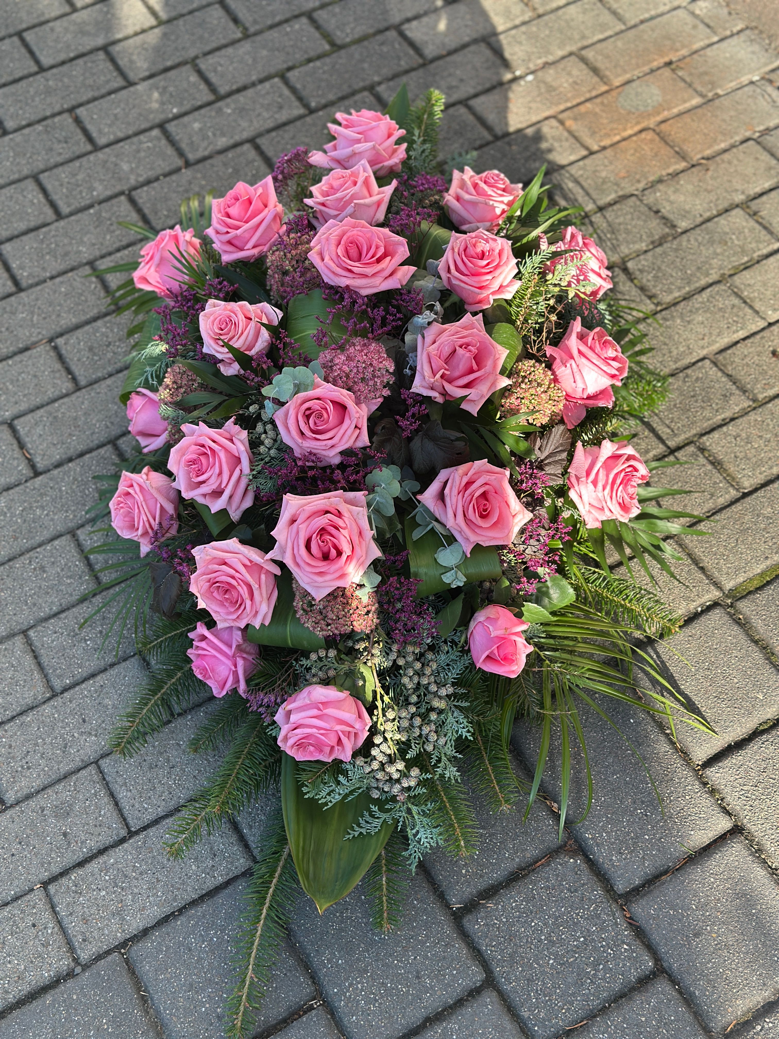Wiązanka pogrzebowa w kształcie łezki z różowych róż, uzupełniona zielonymi dodatkami florystycznymi, ułożona na podkładzie z jodły i florecie z dostępem do wody.