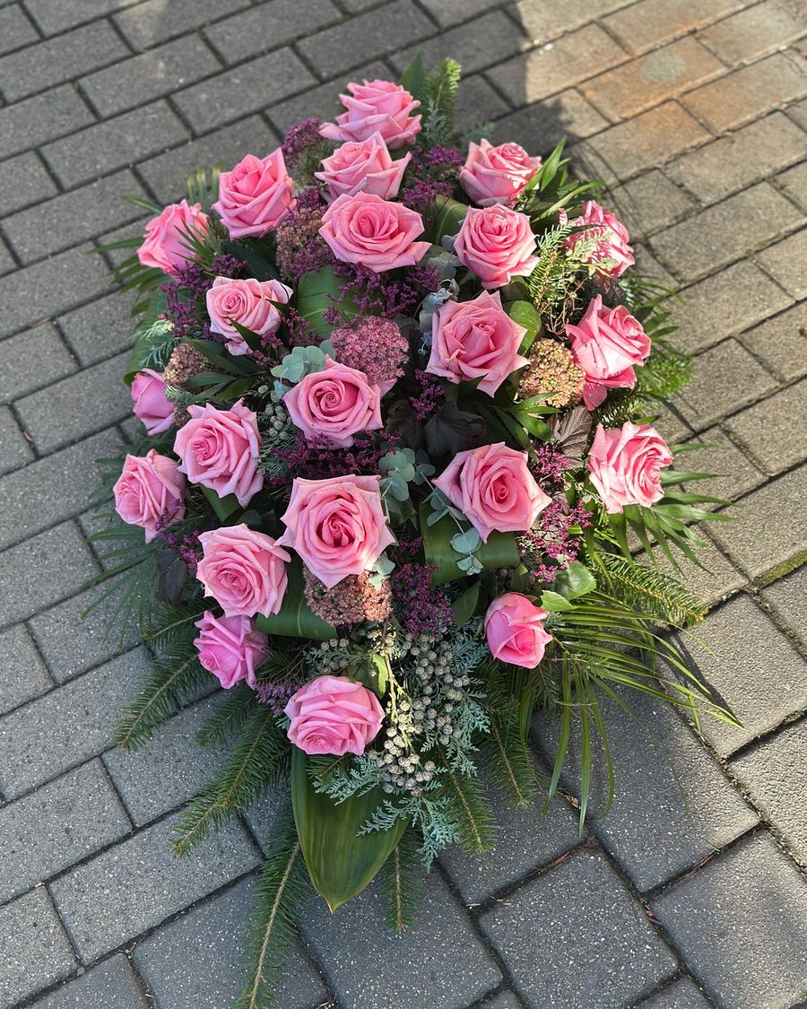 Wiązanka pogrzebowa w kształcie łezki z różowych róż, uzupełniona zielonymi dodatkami florystycznymi, ułożona na podkładzie z jodły i florecie z dostępem do wody.
