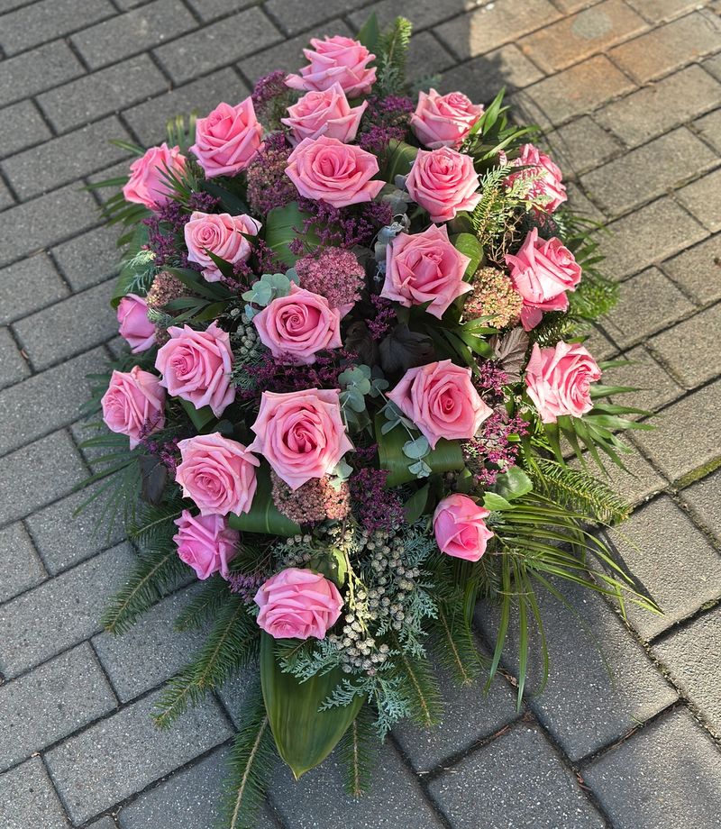 Wiązanka pogrzebowa w kształcie łezki z różowych róż, uzupełniona zielonymi dodatkami florystycznymi, ułożona na podkładzie z jodły i florecie z dostępem do wody.
