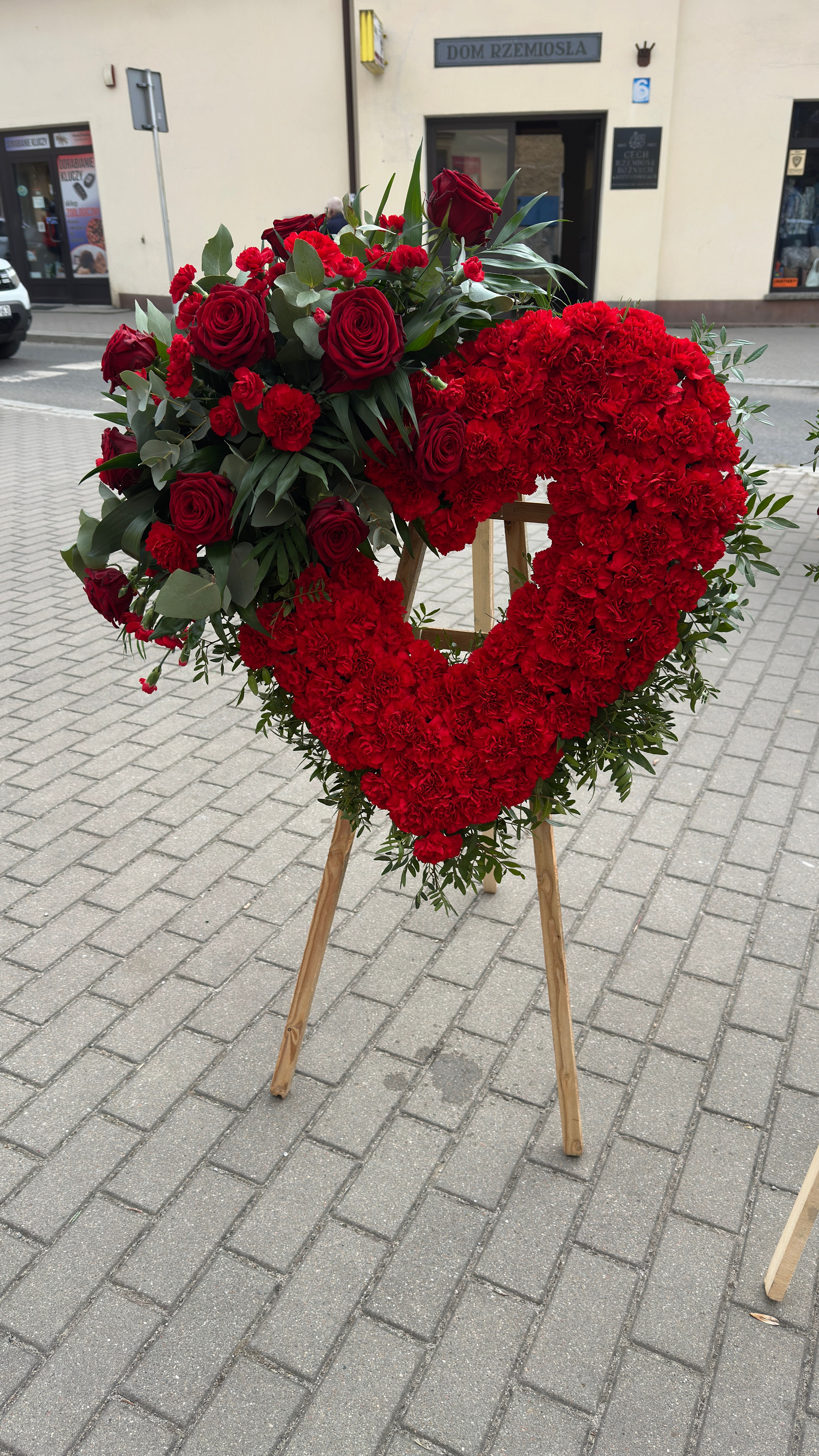 Wieniec w kształcie serca na trzech nogach wykonany z czerwonych goździków i róż.