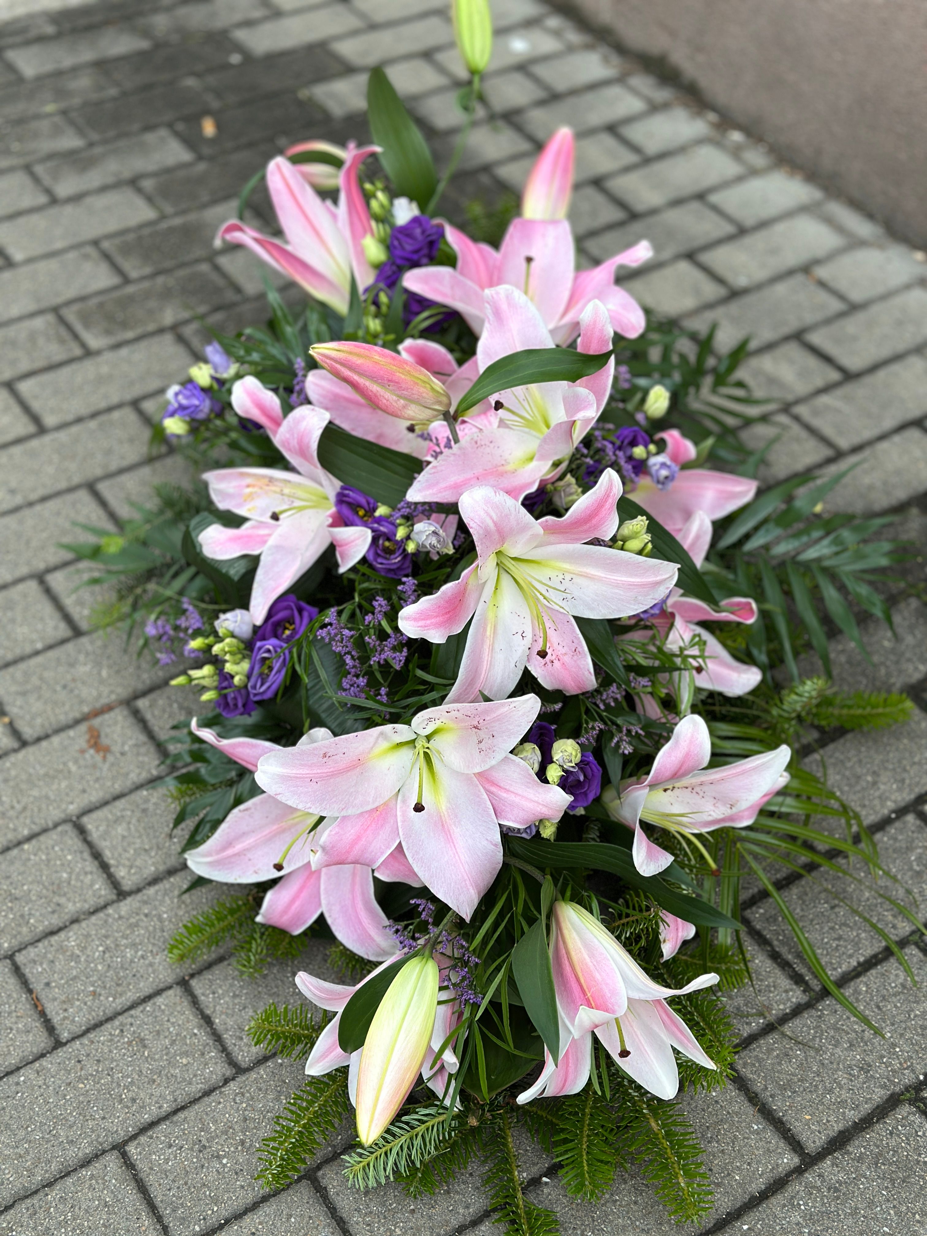 Wiązanka pogrzebowa w kształcie łezki z różowych lilii i fioletowej eustomy, uzupełniona zielonymi dodatkami florystycznymi, ułożona na podkładzie z jodły i florecie z dostępem do wody.