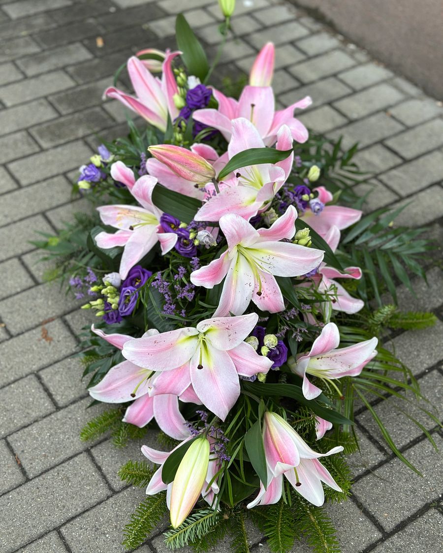 Wiązanka pogrzebowa w kształcie łezki z różowych lilii i fioletowej eustomy, uzupełniona zielonymi dodatkami florystycznymi, ułożona na podkładzie z jodły i florecie z dostępem do wody.