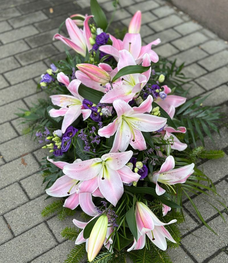 Wiązanka pogrzebowa w kształcie łezki z różowych lilii i fioletowej eustomy, uzupełniona zielonymi dodatkami florystycznymi, ułożona na podkładzie z jodły i florecie z dostępem do wody.