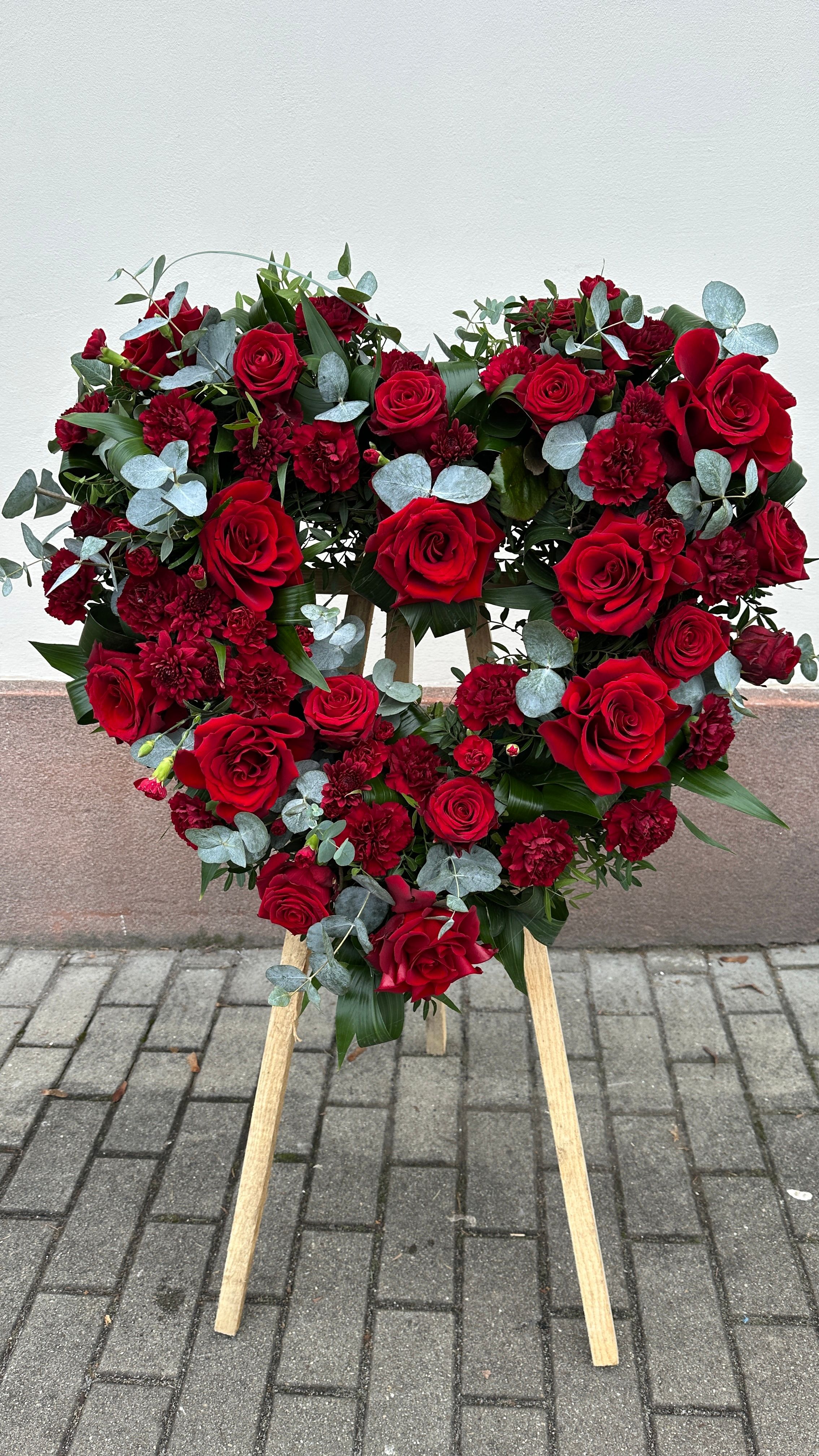 Wieniec pogrzebowy w kształcie serca na trzech nogach wykonany z czerwonych róż, goździka i eukaliptusa.