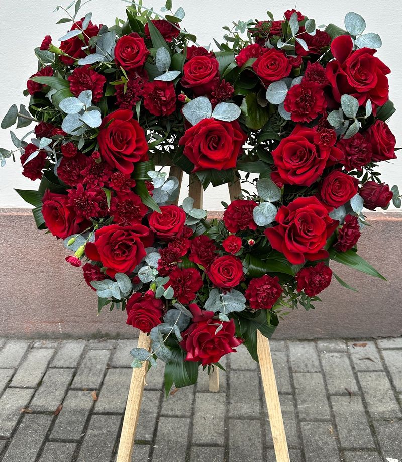 Wieniec pogrzebowy w kształcie serca na trzech nogach wykonany z czerwonych róż, goździka i eukaliptusa.