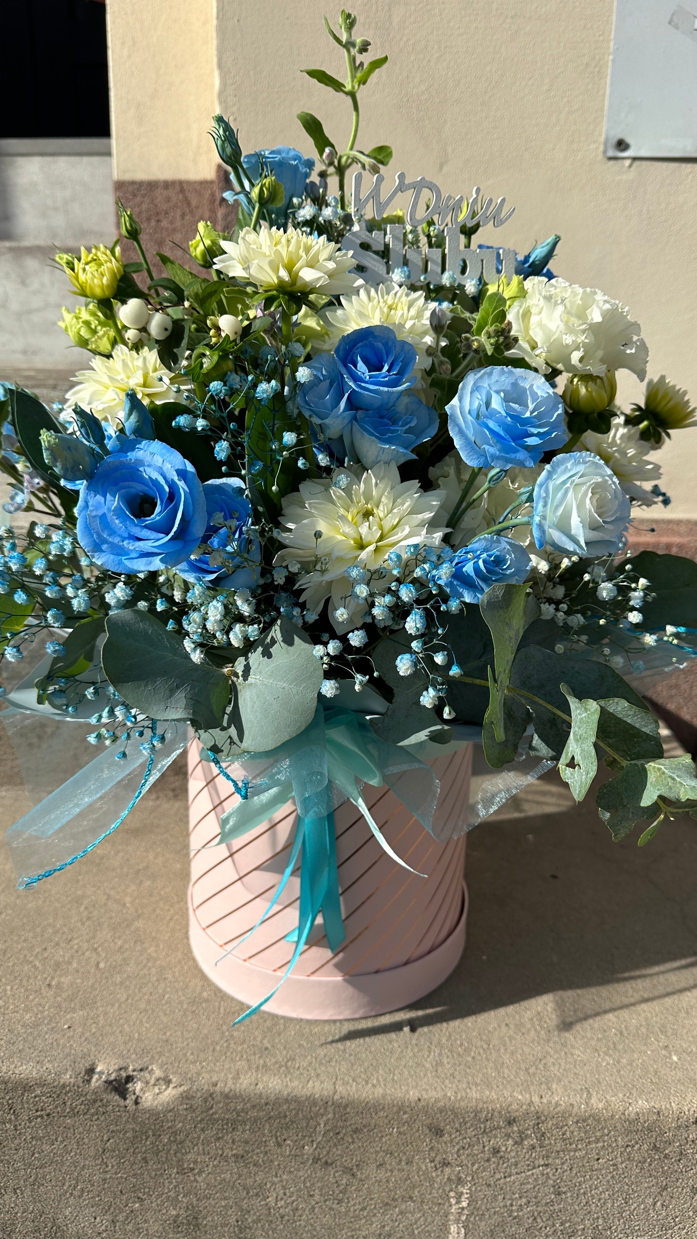 Delikatny flowerbox z niebieską eustomą oraz różnorodnymi kwiatami w kremowych odcieniach to subtelna kompozycja o eleganckim i świeżym charakterze.