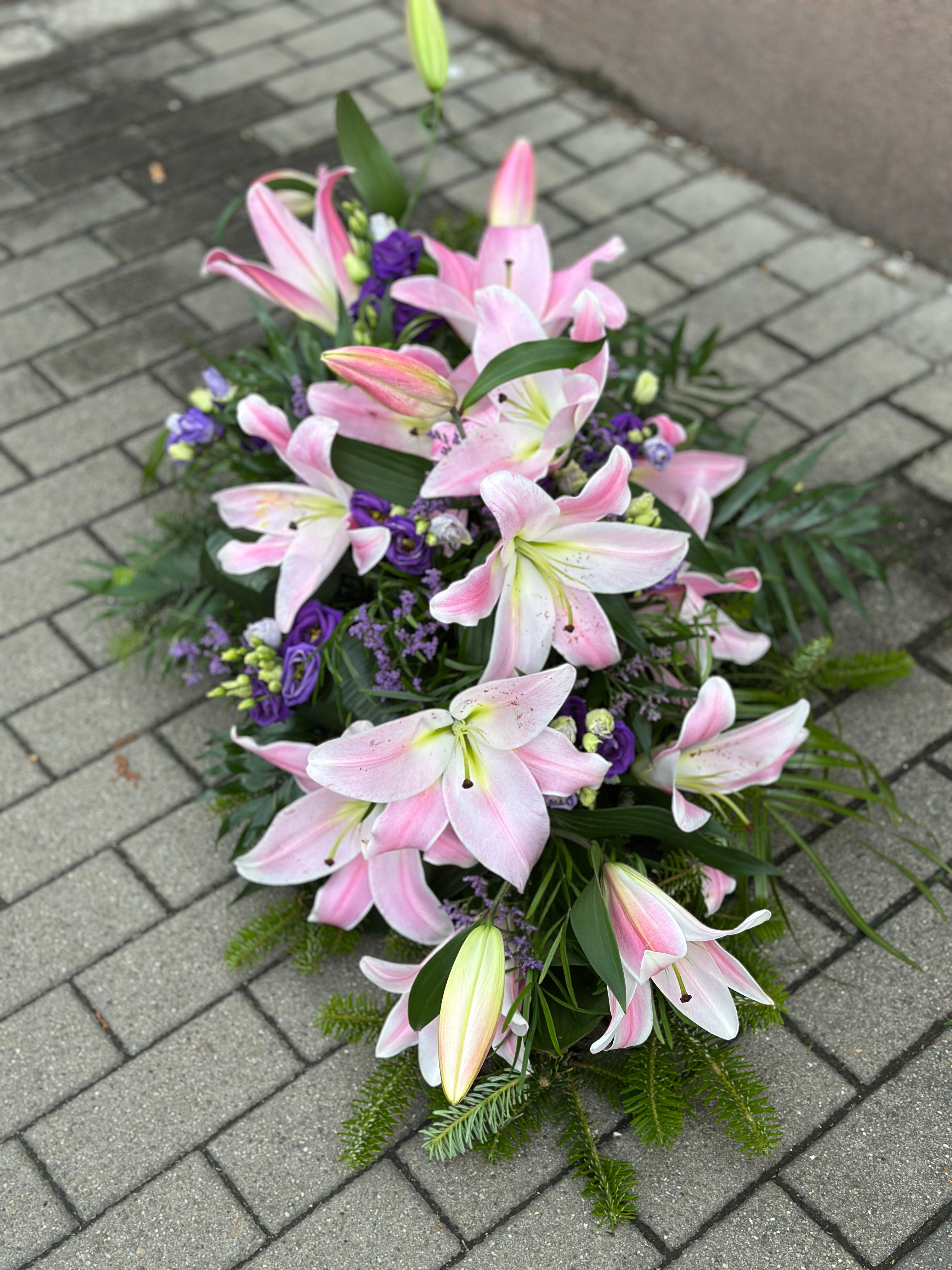 Wiązanka pogrzebowa w kształcie łezki z różowych lilii i fioletowej eustomy, uzupełniona zielonymi dodatkami florystycznymi, ułożona na podkładzie z jodły i florecie z dostępem do wody ujęcie 2.