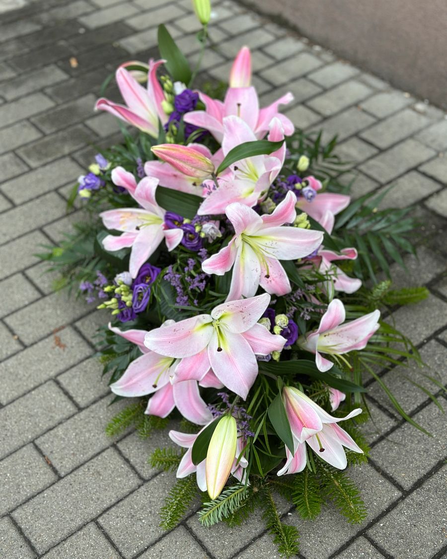 Wiązanka pogrzebowa w kształcie łezki z różowych lilii i fioletowej eustomy, uzupełniona zielonymi dodatkami florystycznymi, ułożona na podkładzie z jodły i florecie z dostępem do wody ujęcie 2.