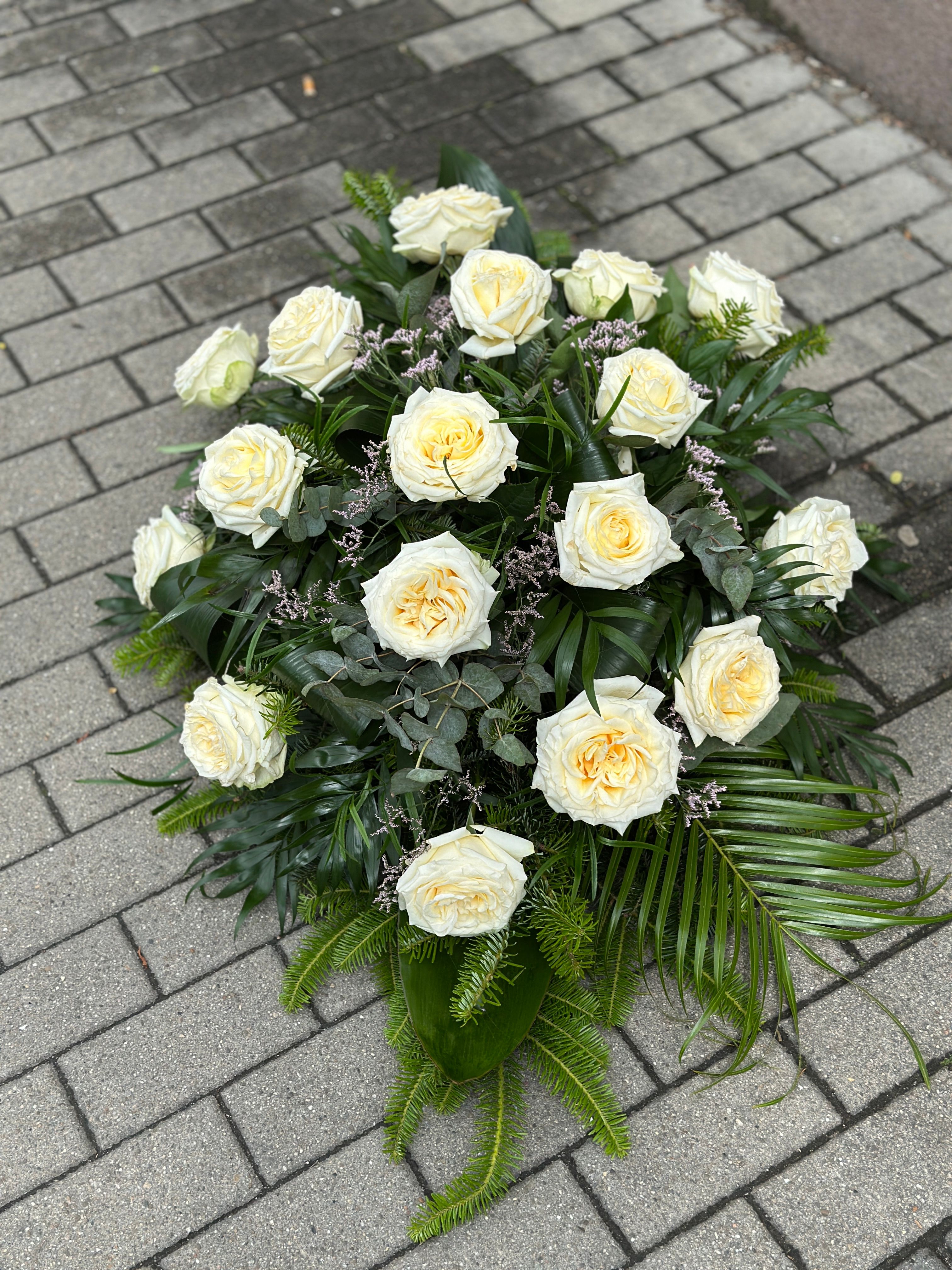 Wiązanka pogrzebowa w kształcie łezki z kremowych róż, uzupełniona zielonymi dodatkami florystycznymi, ułożona na podkładzie z jodły i florecie z dostępem do wody.