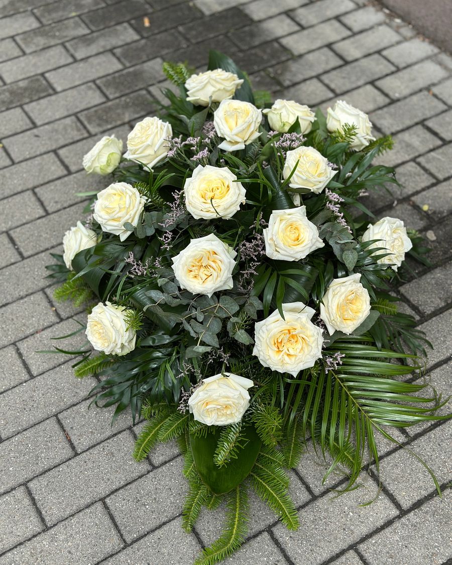 Wiązanka pogrzebowa w kształcie łezki z kremowych róż, uzupełniona zielonymi dodatkami florystycznymi, ułożona na podkładzie z jodły i florecie z dostępem do wody.