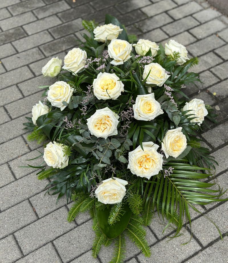 Wiązanka pogrzebowa w kształcie łezki z kremowych róż, uzupełniona zielonymi dodatkami florystycznymi, ułożona na podkładzie z jodły i florecie z dostępem do wody.