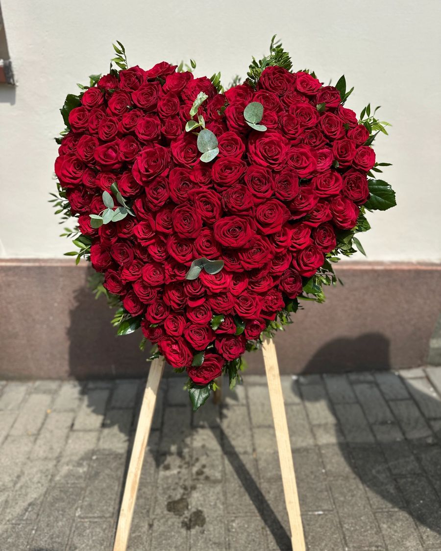 Wieniec pogrzebowy w kształcie serca na trzech nogach z czerwonych róż z zielonymi dodatkami.