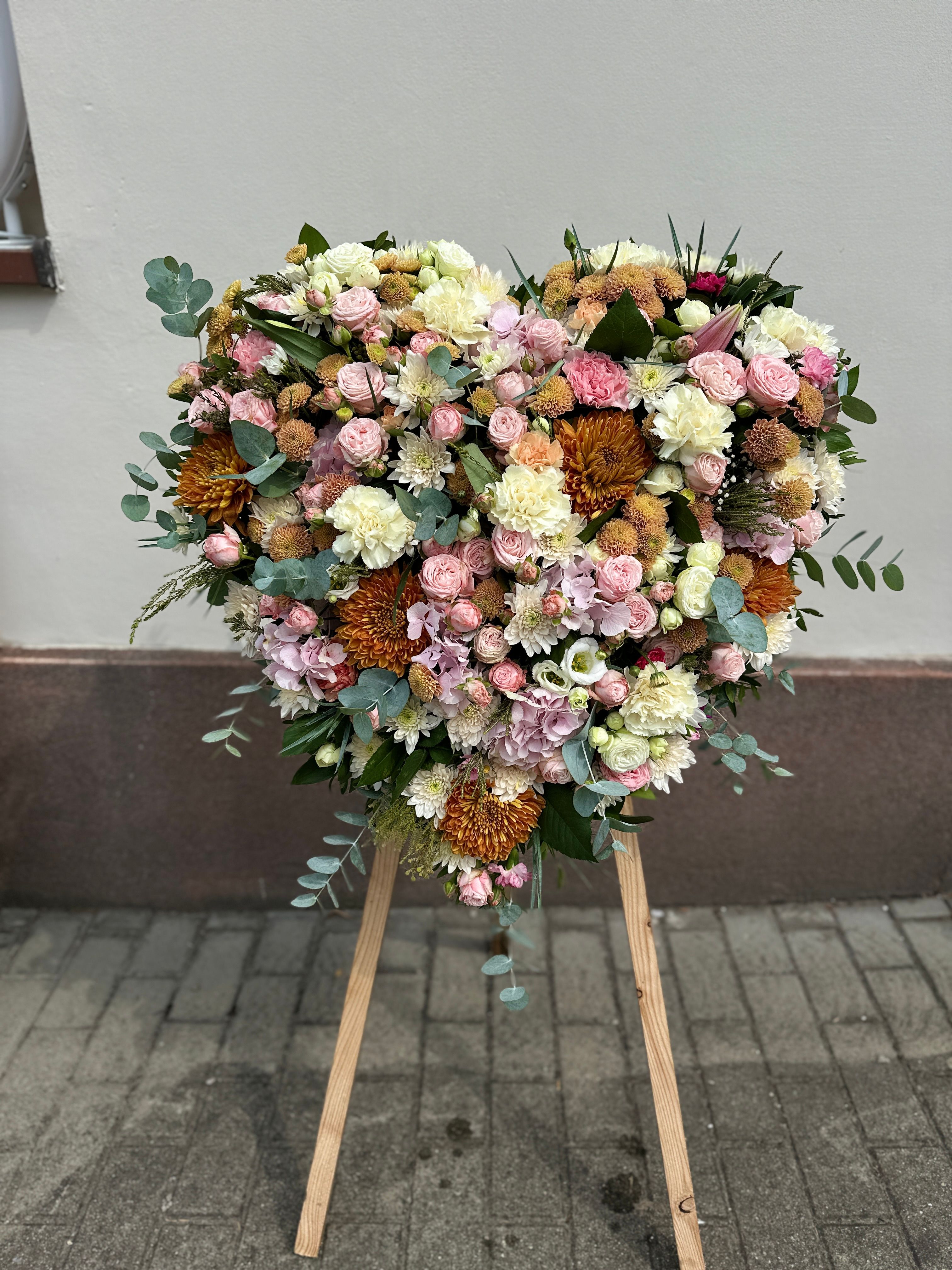 Wieniec pogrzebowy w kształcie serca na trzech nogach wykonany z różnorodnych kwiatów w odcieniach pastelowych różu i kremu.