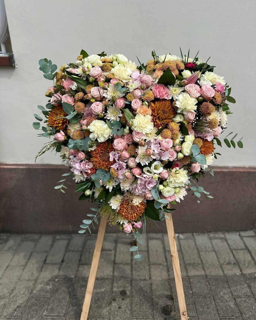 Wieniec pogrzebowy w kształcie serca na trzech nogach wykonany z różnorodnych kwiatów w odcieniach pastelowych różu i kremu.