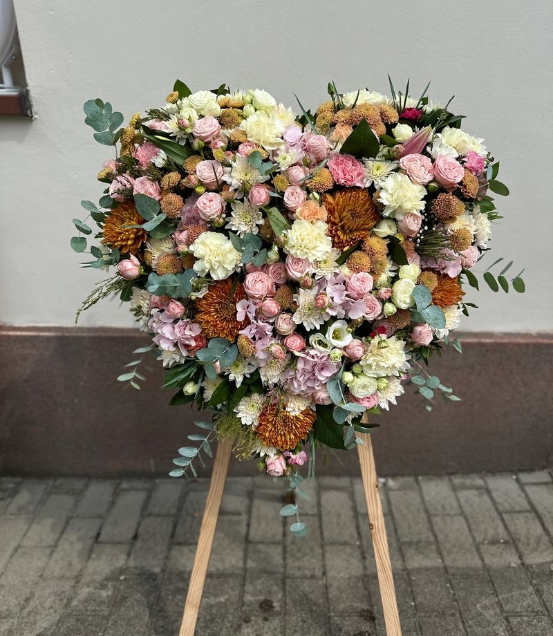 Wieniec pogrzebowy w kształcie serca na trzech nogach wykonany z różnorodnych kwiatów w odcieniach pastelowych różu i kremu.