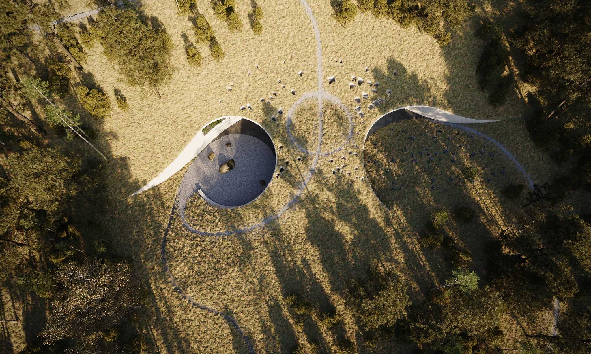 Aerial view of two large, curved architectural installations set within a vast, dry grassy field. One structure is a circular depression with a grey gravel floor and scattered rocks, while the other is an open crescent-shaped wall enclosing a grassy area with dark accents. Long tree shadows stretch across the landscape, which features distinctive winding paths and numerous small boulders between the modern structures.
