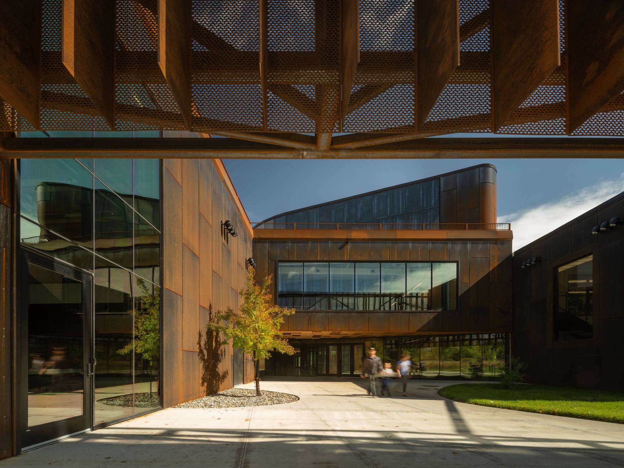 Modern architectural complex featuring weathered Corten steel panels, large glass windows, and an intricate perforated metal canopy overhead. A bright outdoor courtyard with blurred figures walking leads to the building entrance.