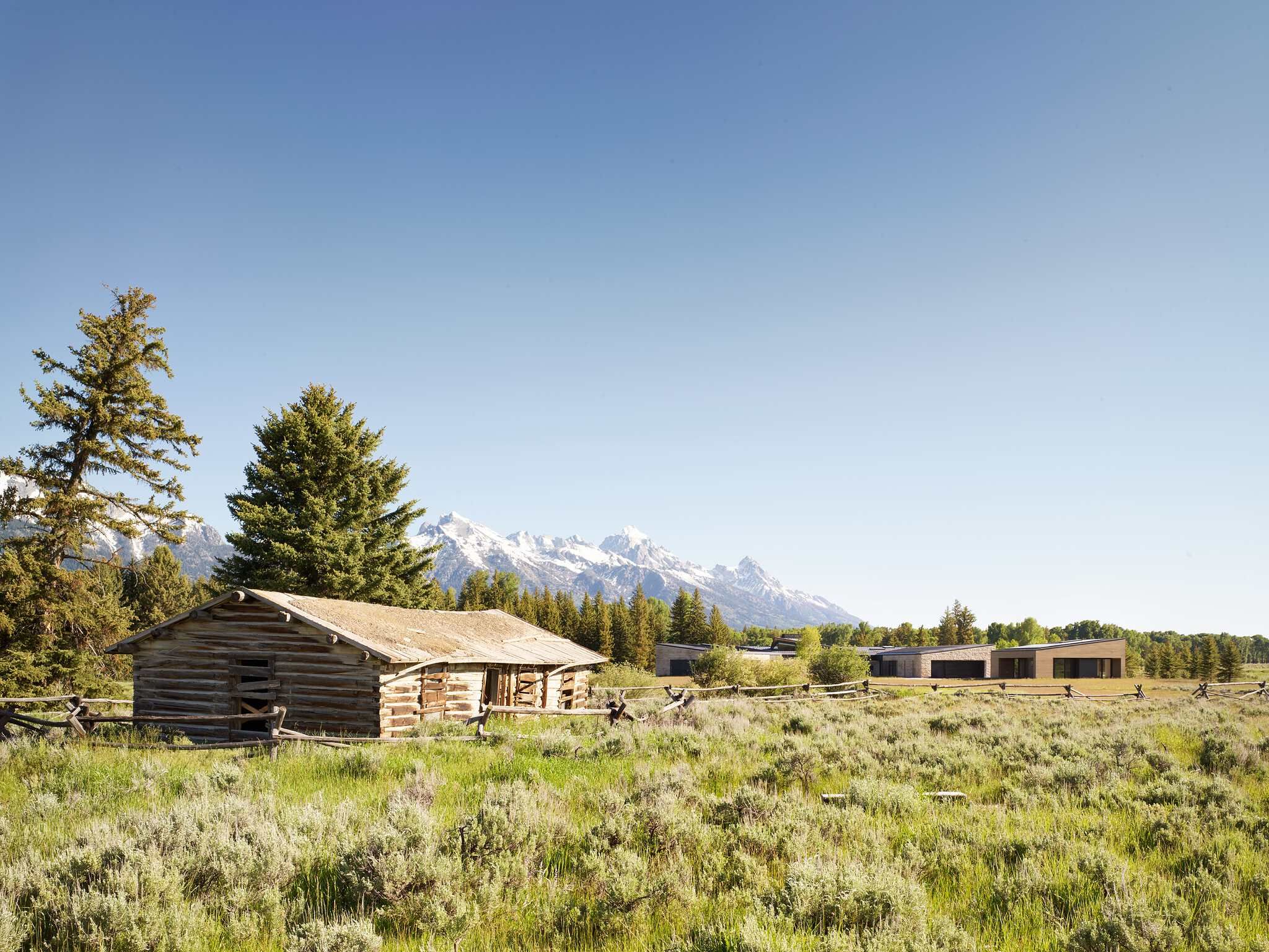 Weathered log cabin and split rail fence in a vast sagebrush field, with modern buildings, pine trees, and the snow-capped Grand Teton Range in the distance under a clear blue sky.