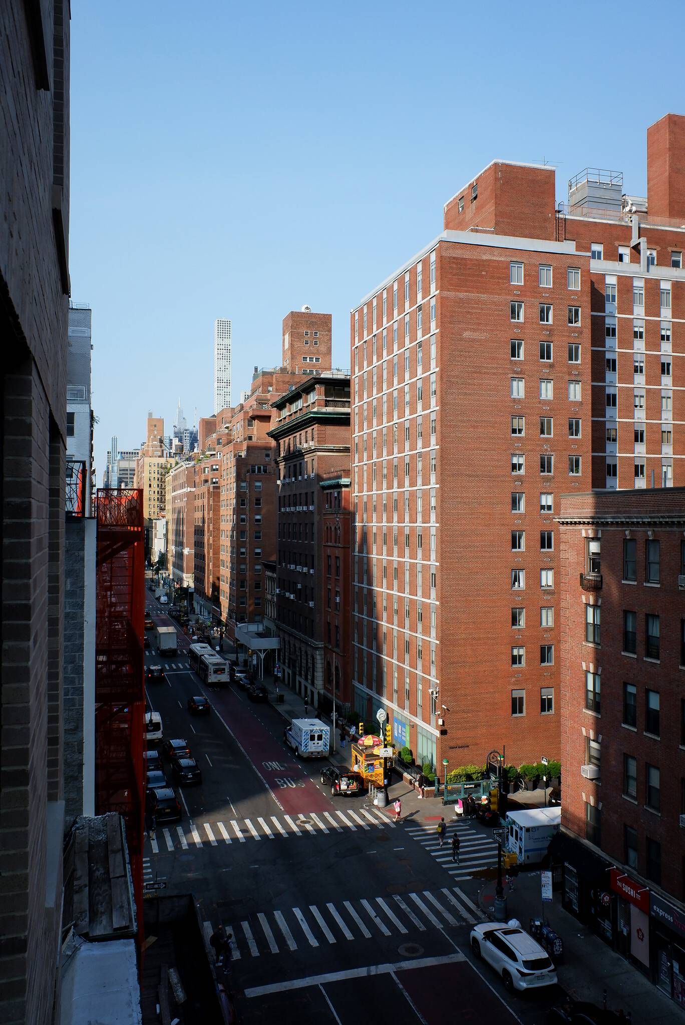 Elevated view of a bustling city street lined with multi-story red brick buildings, featuring cars, a bus, pedestrians, and street vendors. A tall, slender white skyscraper rises in the distant background, with sunlight casting long shadows across the road.