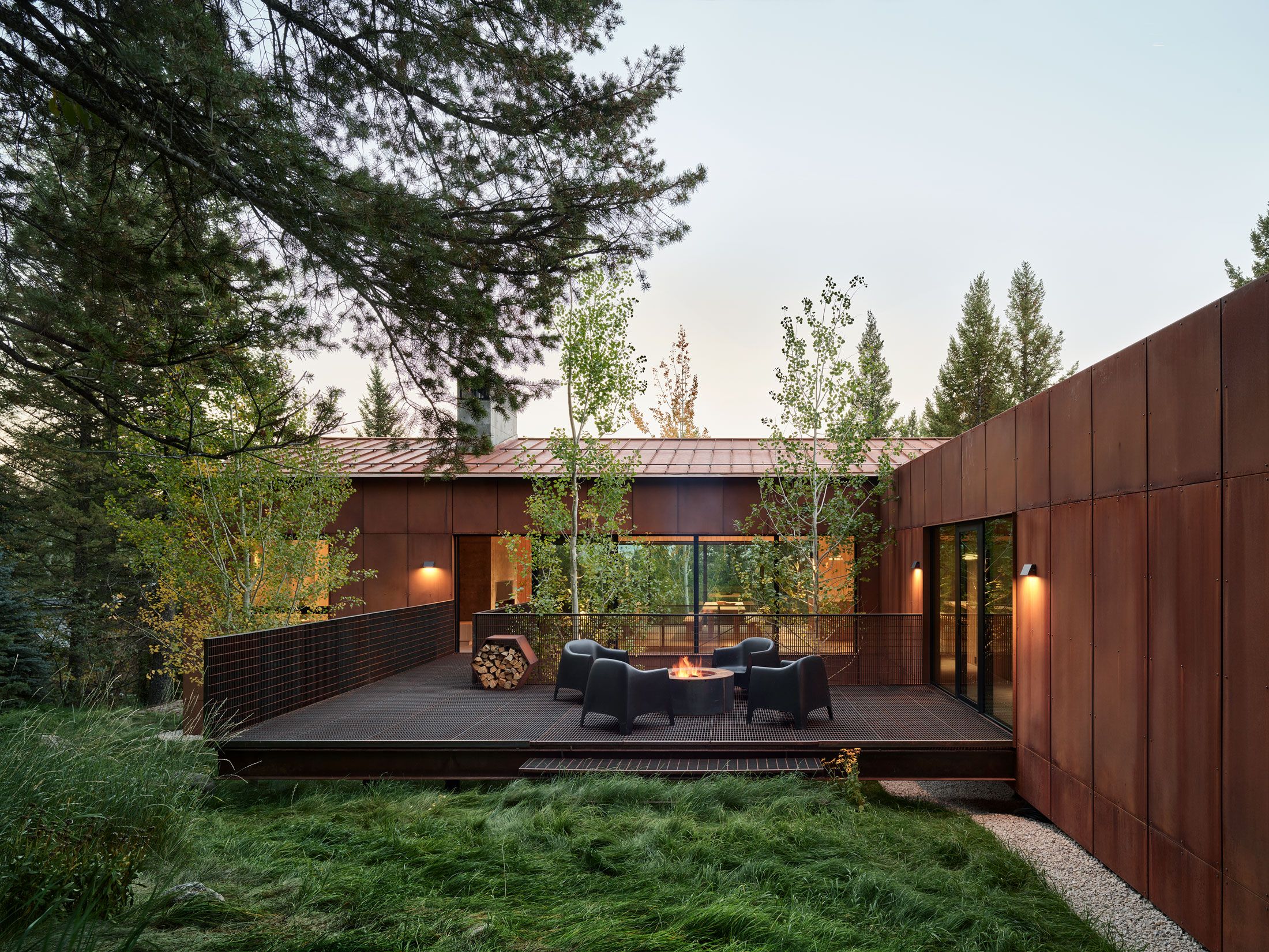 Modern Corten steel house with an outdoor metal deck, fire pit, and seating, surrounded by tall aspens, pines, and natural grasses at twilight.