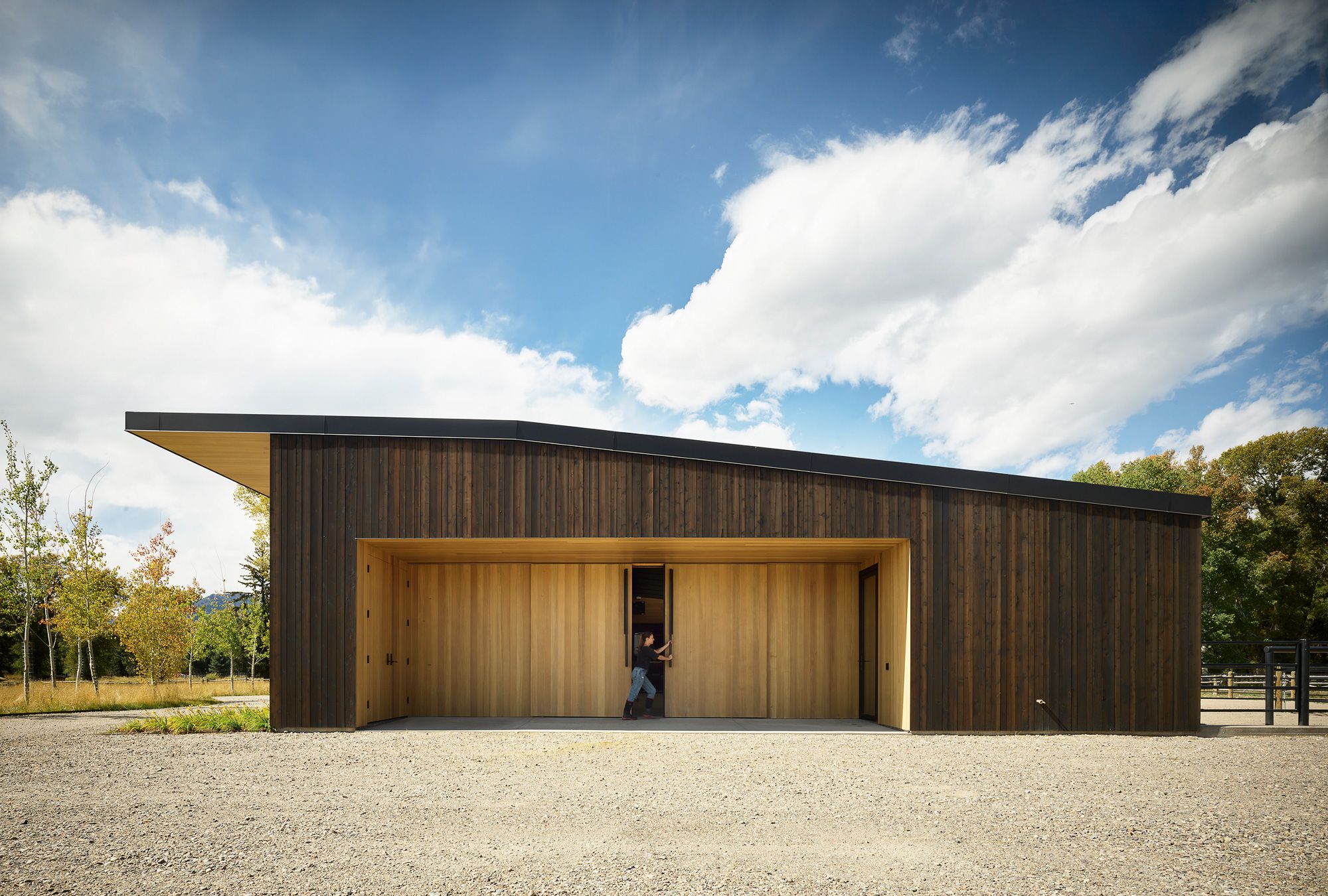 Modern stable with dark vertical wood siding and a black shed roof. A person opens one of the large, light wood sliding doors in a recessed entrance, facing a gravel foreground, autumnal trees, and a blue sky with white clouds.