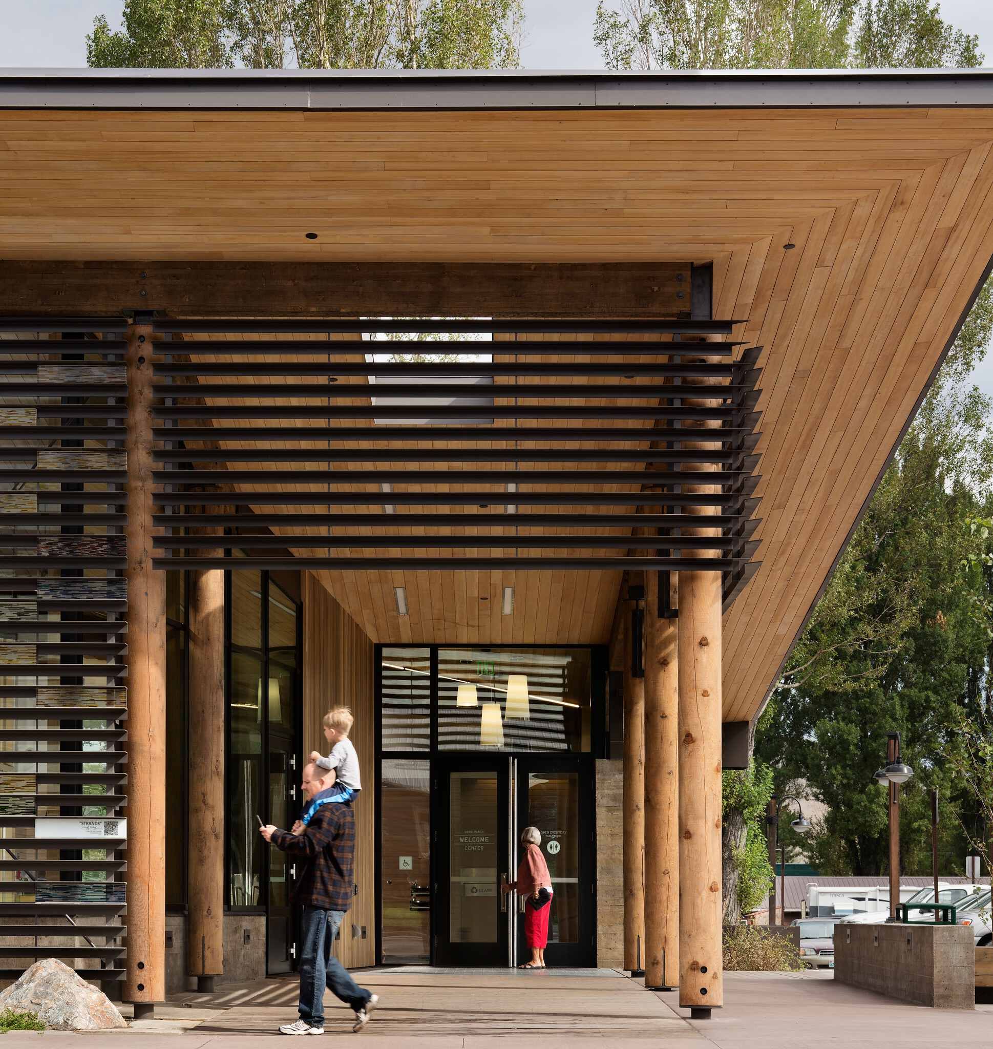 Modern building entrance featuring a slatted wood ceiling, large log columns, and horizontal metal sun shades. A man carries a child on his shoulders as a woman enters the glass doors of the Welcome Center.