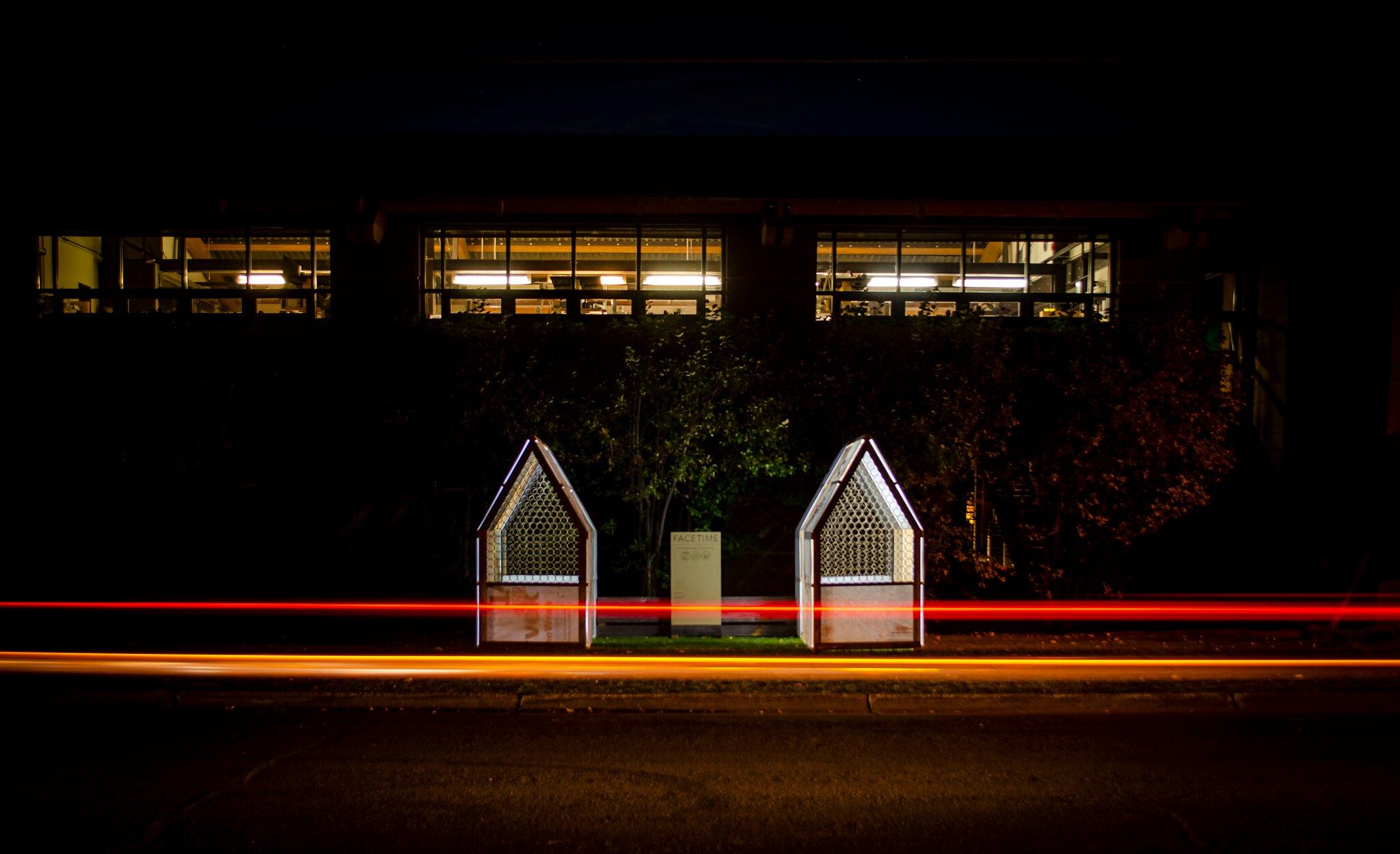 Urban night scene featuring vibrant red and orange long-exposure light trails above a dark street. Two house-shaped art installations with honeycomb-patterned fronts flank a central FACETIME sign, positioned in front of a building with brightly lit windows.