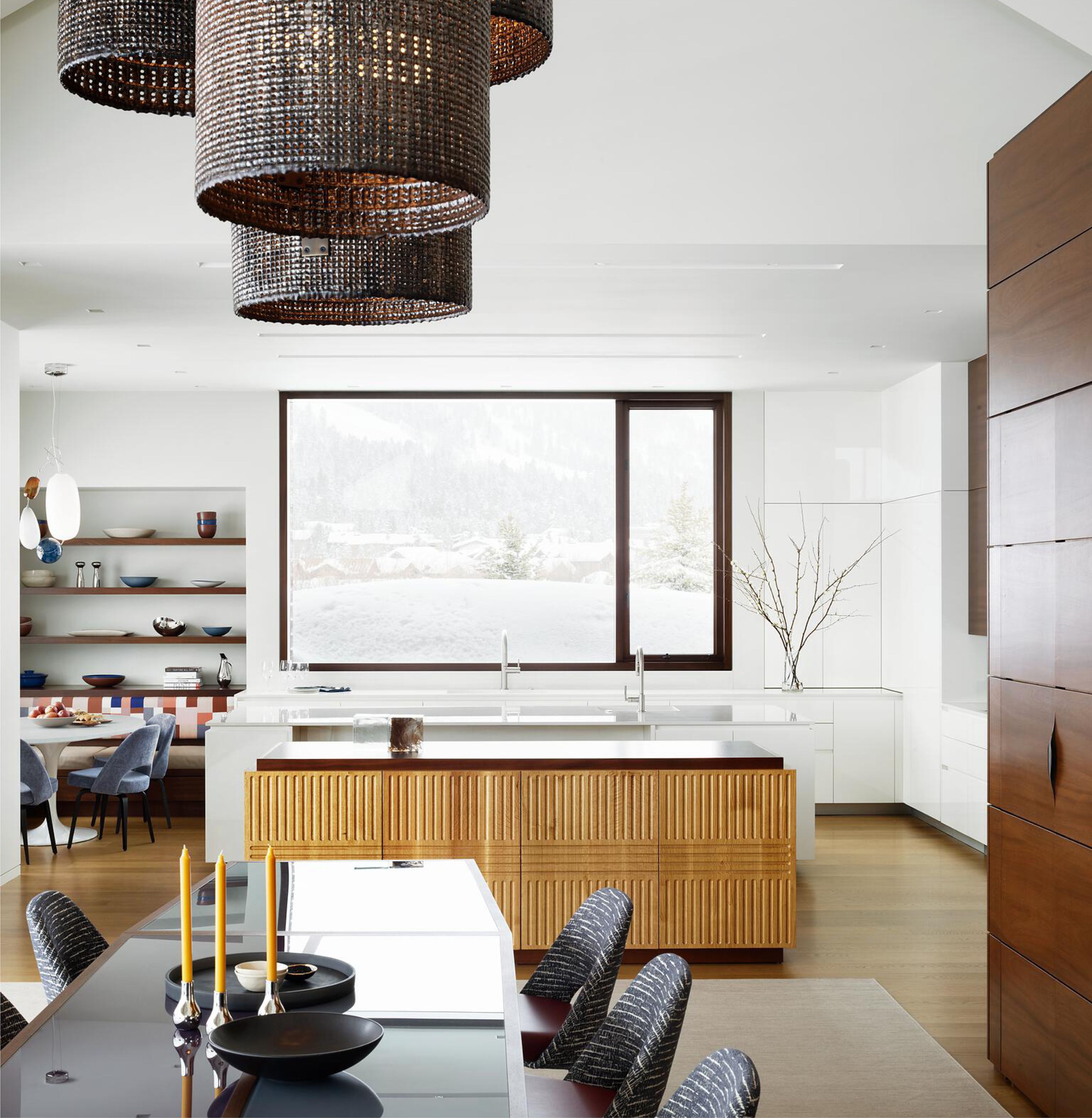 Contemporary kitchen and dining room featuring large dark woven pendant lights, a fluted wooden island, and a dining table set with yellow candles in the foreground, with a snowy mountain landscape visible through a large window.