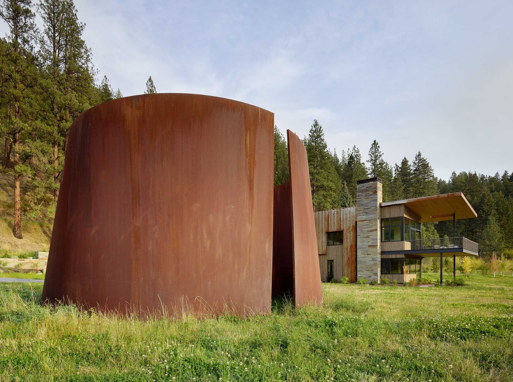 A modern house with a stone chimney, wood siding, and a cantilevered deck stands behind a large, curved Corten steel sculpture in a grassy meadow, backed by a dense pine forest and hillside.