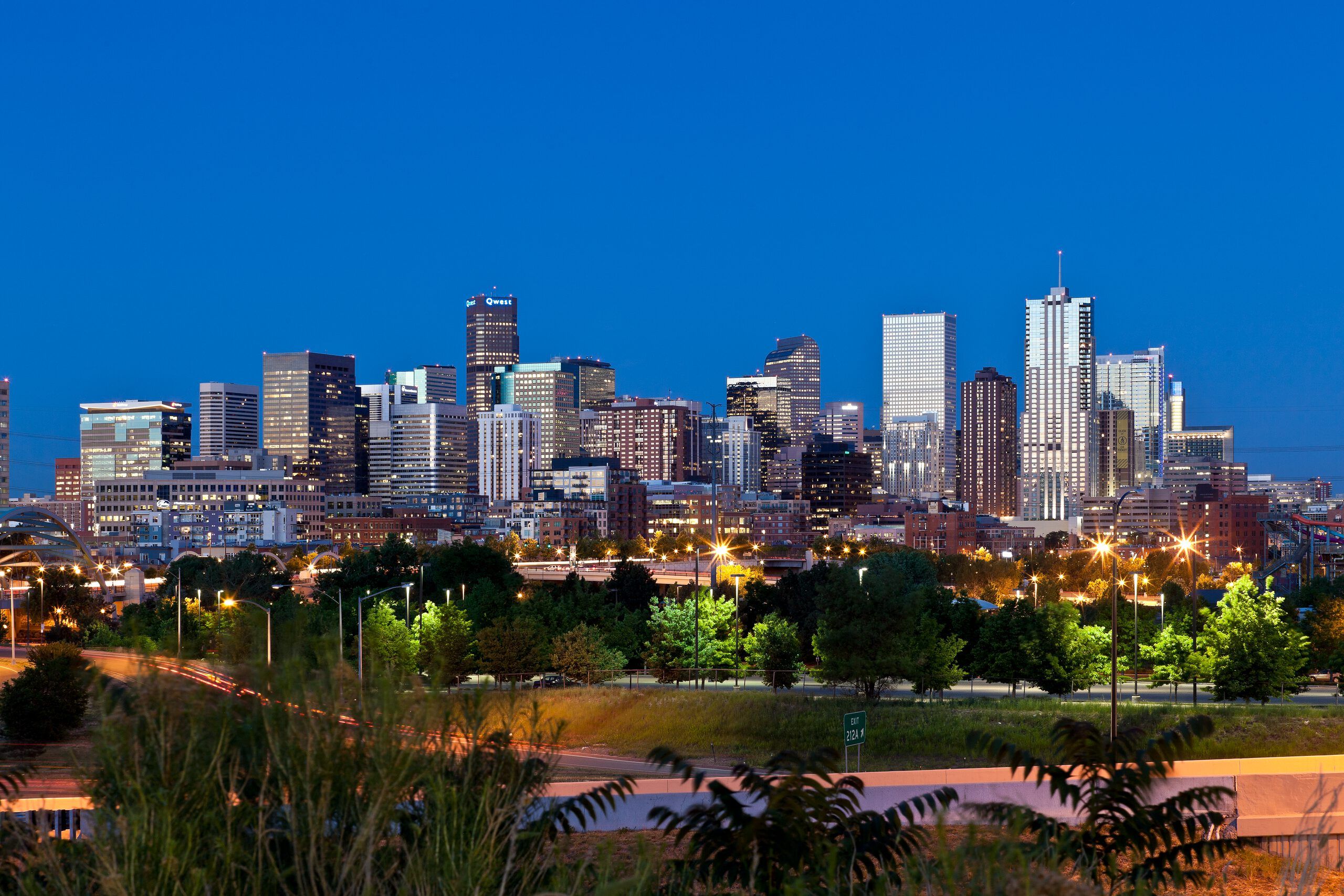 Denver, Colorado, city skyline at dusk with illuminated skyscrapers, a highway with car light trails, and lush trees in the foreground.