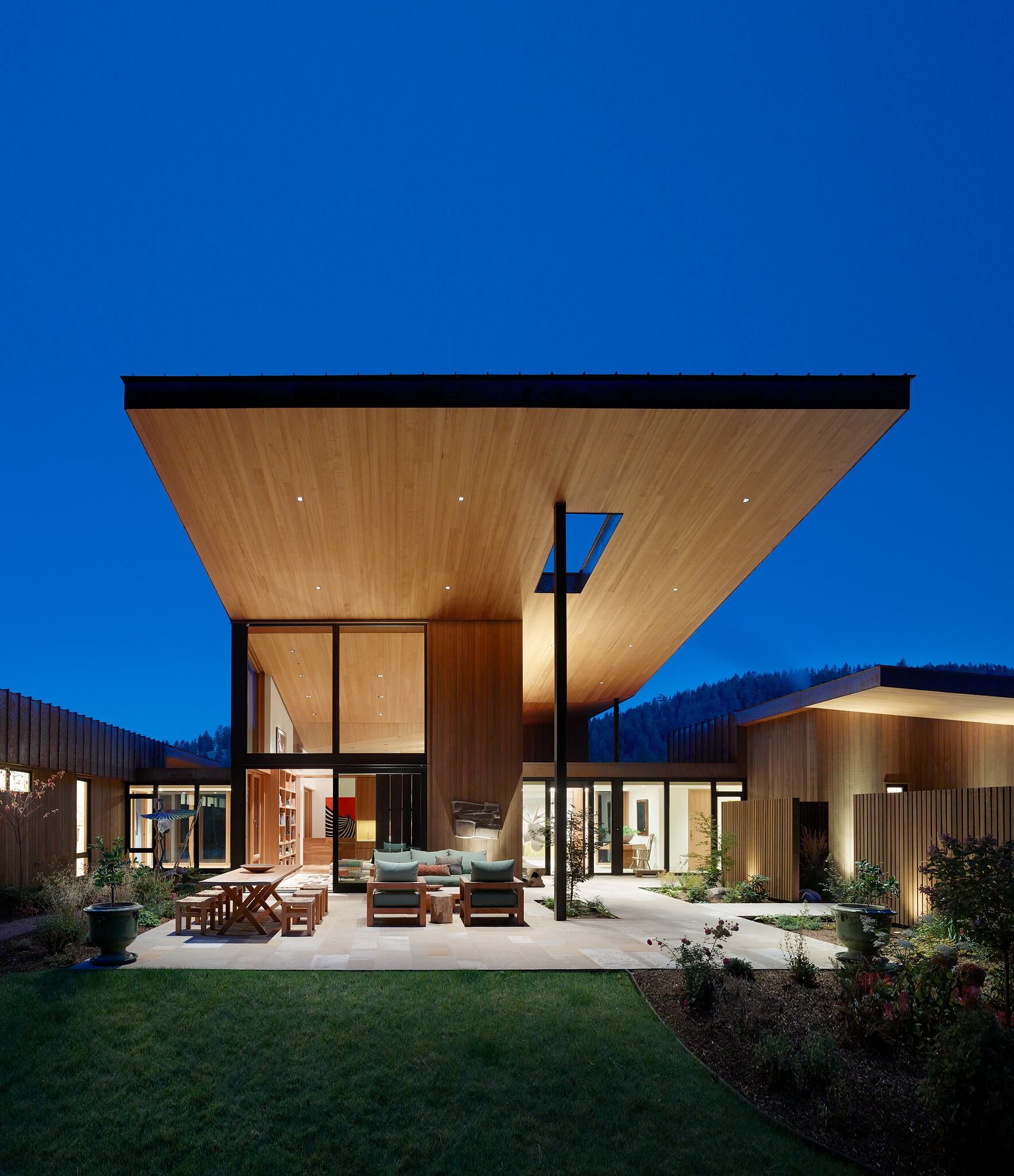 A striking contemporary luxury home illuminated under a deep blue twilight sky. The residence features an expansive cantilevered wooden roof, supported by a dark column and featuring a rectangular skylight, extending over a paved patio. Floor-to-ceiling glass windows reveal warm interior lighting. The patio includes modern outdoor dining and lounge furniture, bordered by a manicured lawn and landscaped gardens. Dark, tree-covered hills are visible in the background.