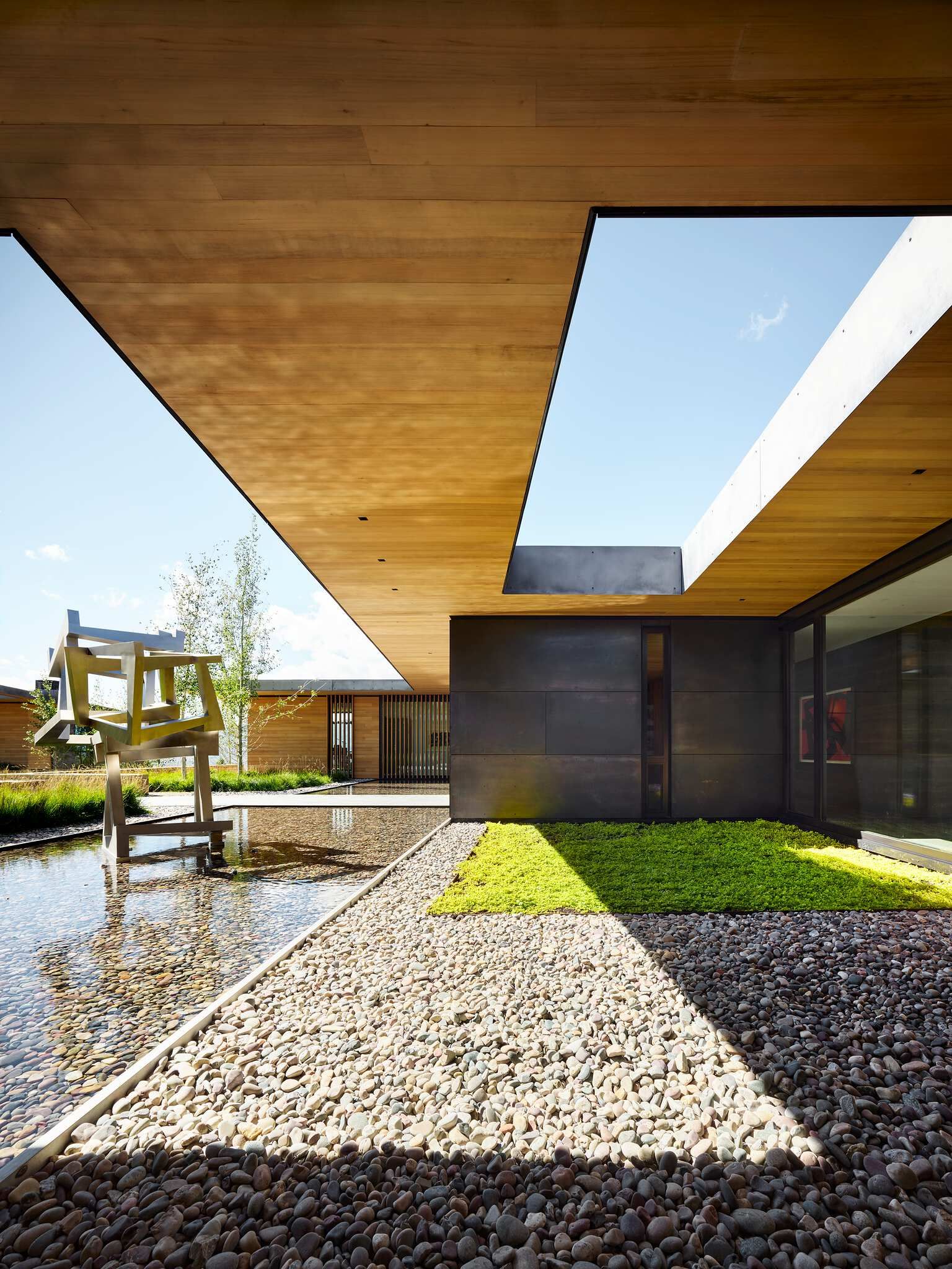 Modern architectural courtyard with a large wooden overhead structure open to the blue sky, featuring an abstract metal sculpture in a reflecting pool, pebble landscaping, and dark metal building facades with tall windows.