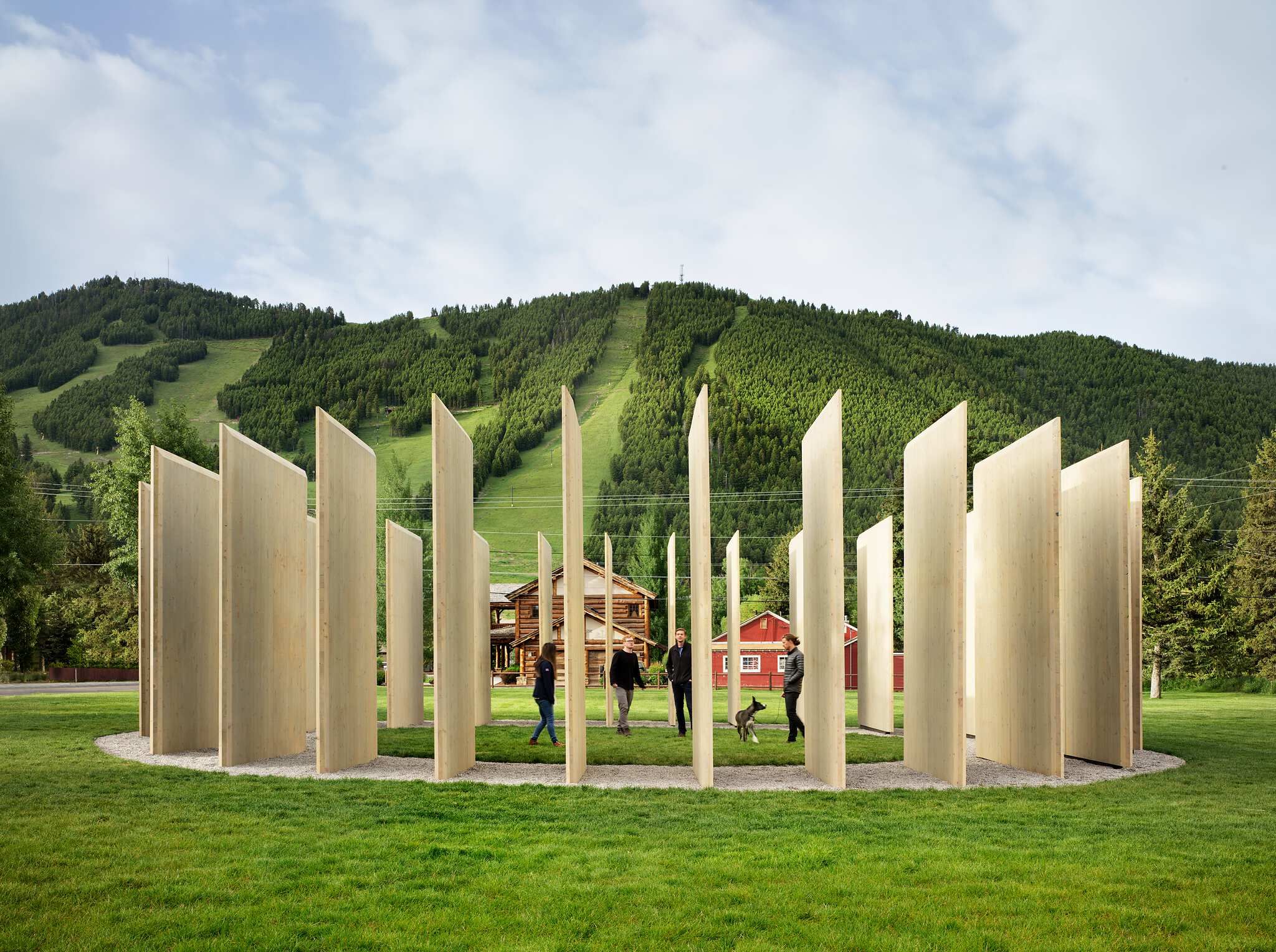 A circular outdoor art installation made of tall, light-colored wooden panels on a gravel path in a grassy area, with people and a dog exploring it. A forested mountain with ski slopes, a rustic log cabin, and a red barn are visible in the background under a cloudy sky.