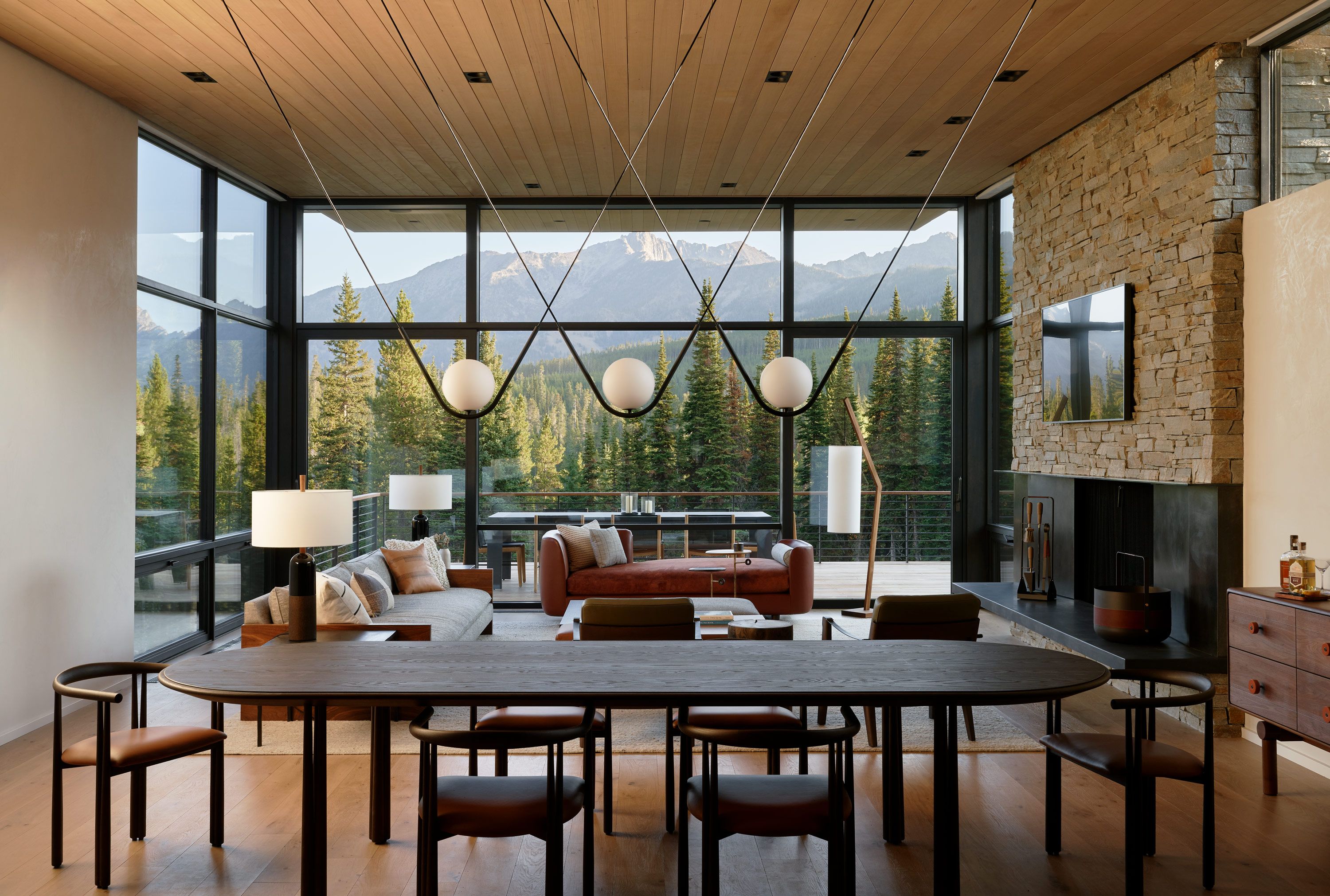 Spacious modern living and dining room with panoramic floor-to-ceiling windows showcasing a stunning mountain and evergreen forest view. A striking multi-globe chandelier hangs above a dark oval dining table. The adjacent living area features a grey sofa, an orange velvet chaise lounge, and a prominent stone fireplace with a mounted TV, all under a warm wooden paneled ceiling and hardwood floors.