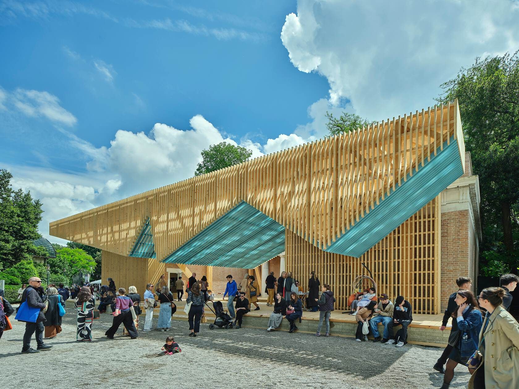 A striking modern architectural pavilion featuring a slatted wooden facade and angular blue panels, extending over an older brick structure. Many visitors are gathered on the gravel ground and a raised platform in front of the building under a bright blue sky with white clouds.